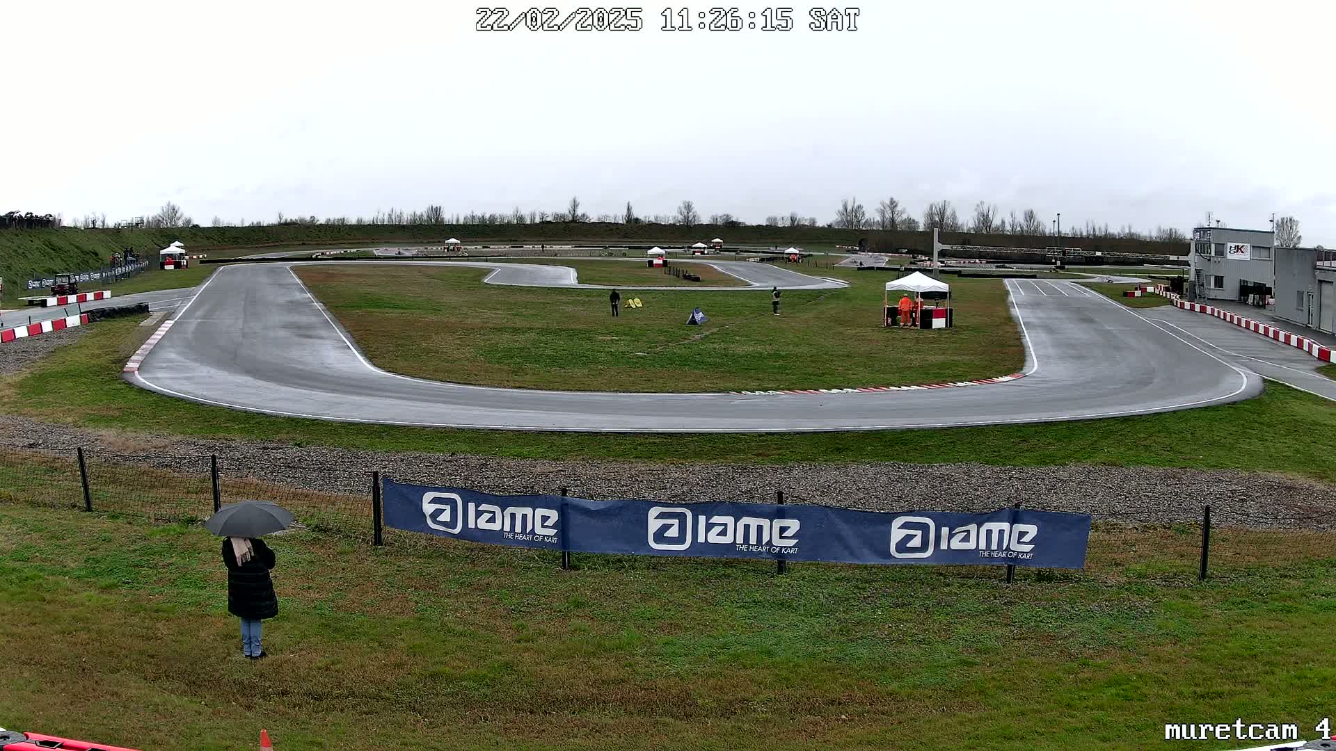 A go-kart track on an overcast day with a few people present and a person with an umbrella standing near a fence.