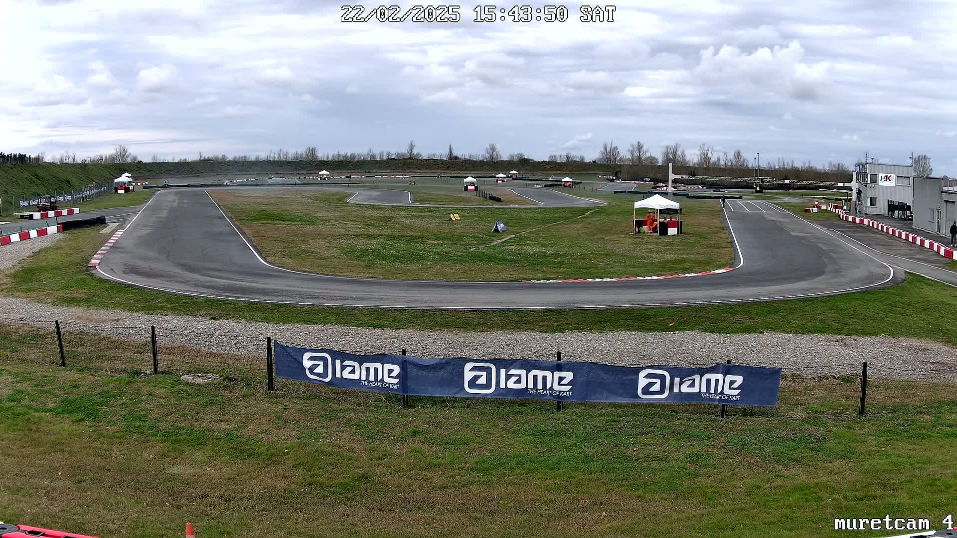 An outdoor go-kart track on an overcast day with some patchy grass and a few people visible near tents.