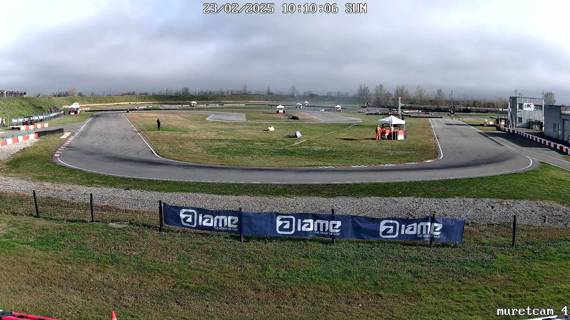 A mostly sunny day at an outdoor go-kart track with several karts visible on the course and spectators watching from the sidelines.