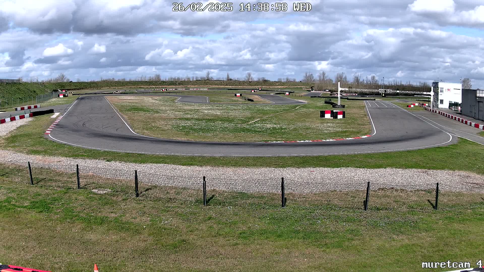 An outdoor go-kart track with a partly cloudy sky is shown, featuring a paved course surrounded by grass and fencing.