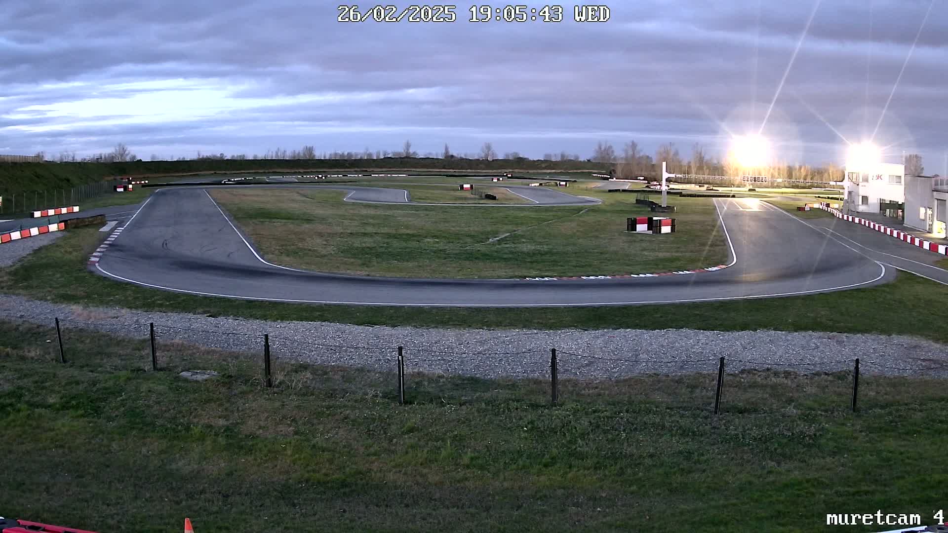 An empty asphalt go-kart track under a cloudy twilight sky is illuminated by bright lights.