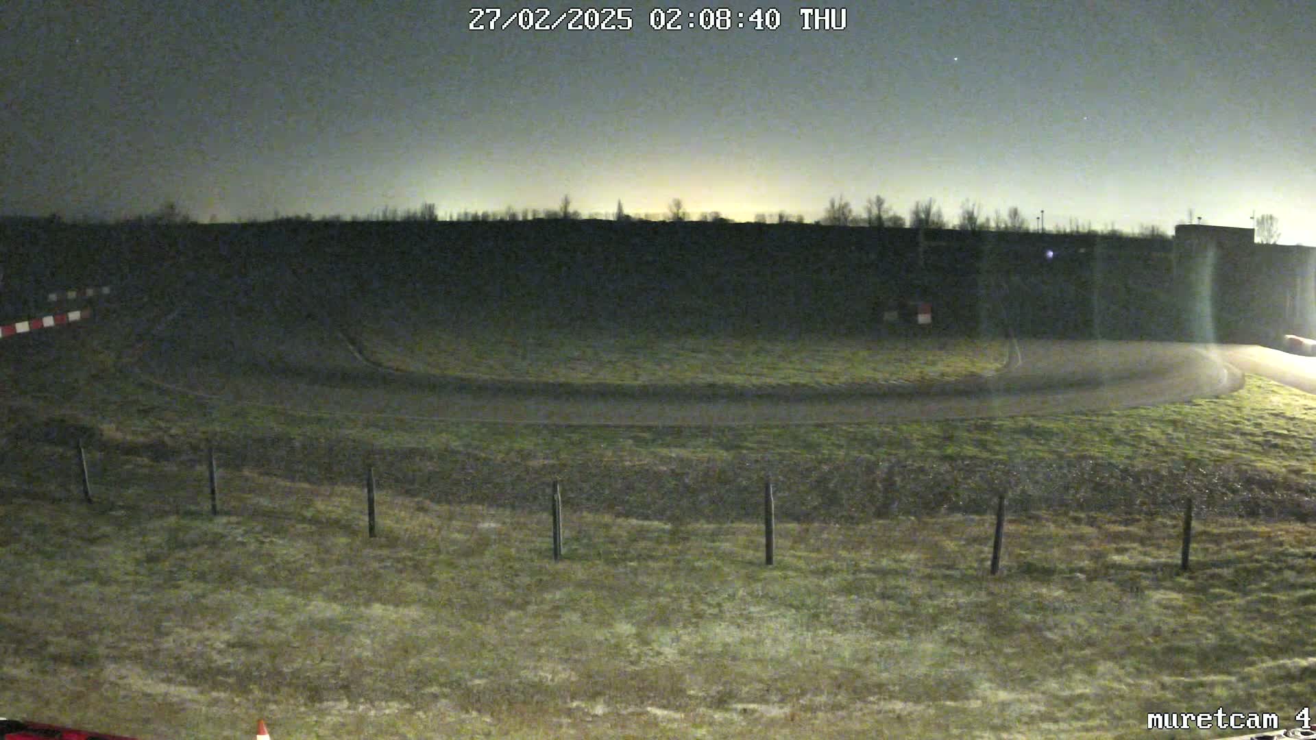 A nighttime, dimly lit view of a paved racetrack curving to the right, surrounded by short, grassy fields under a dark sky.