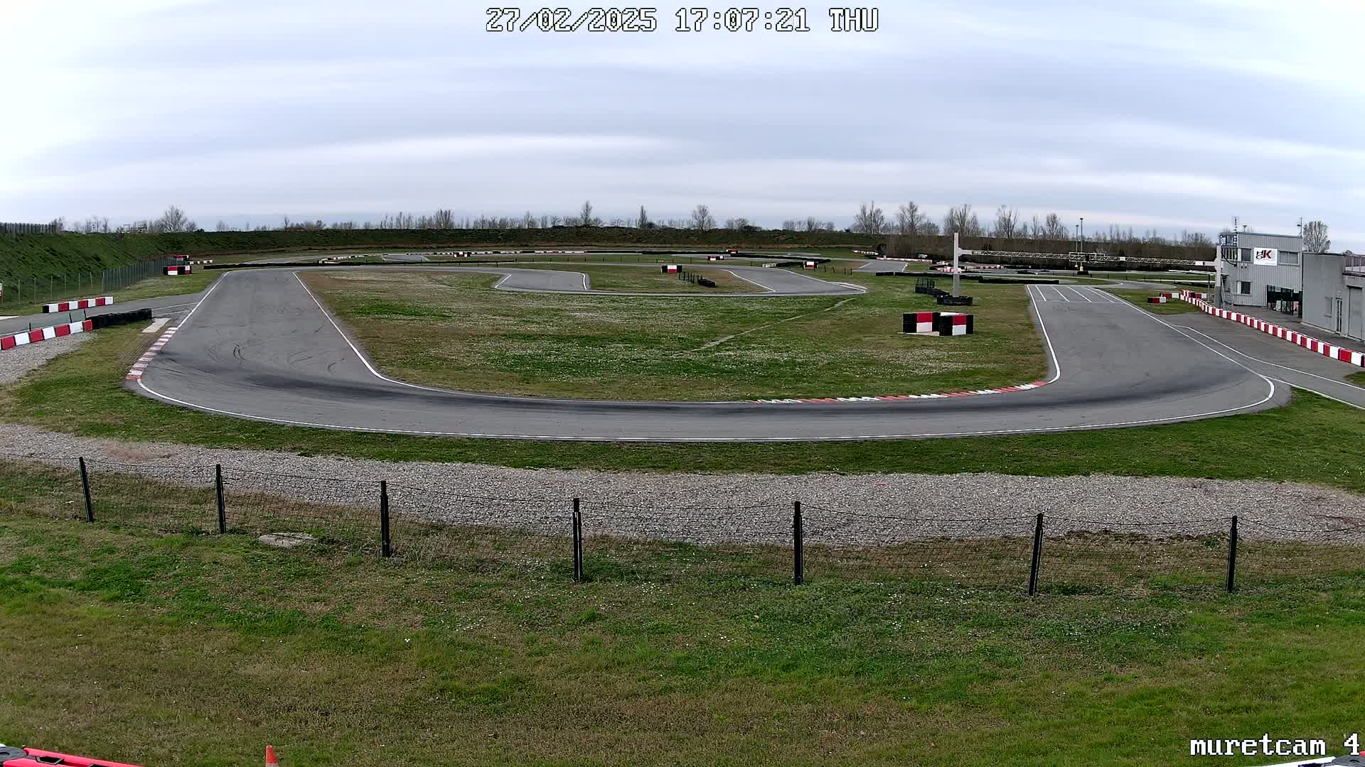 An outdoor go-kart track with a gray asphalt surface and grassy areas under an overcast sky.