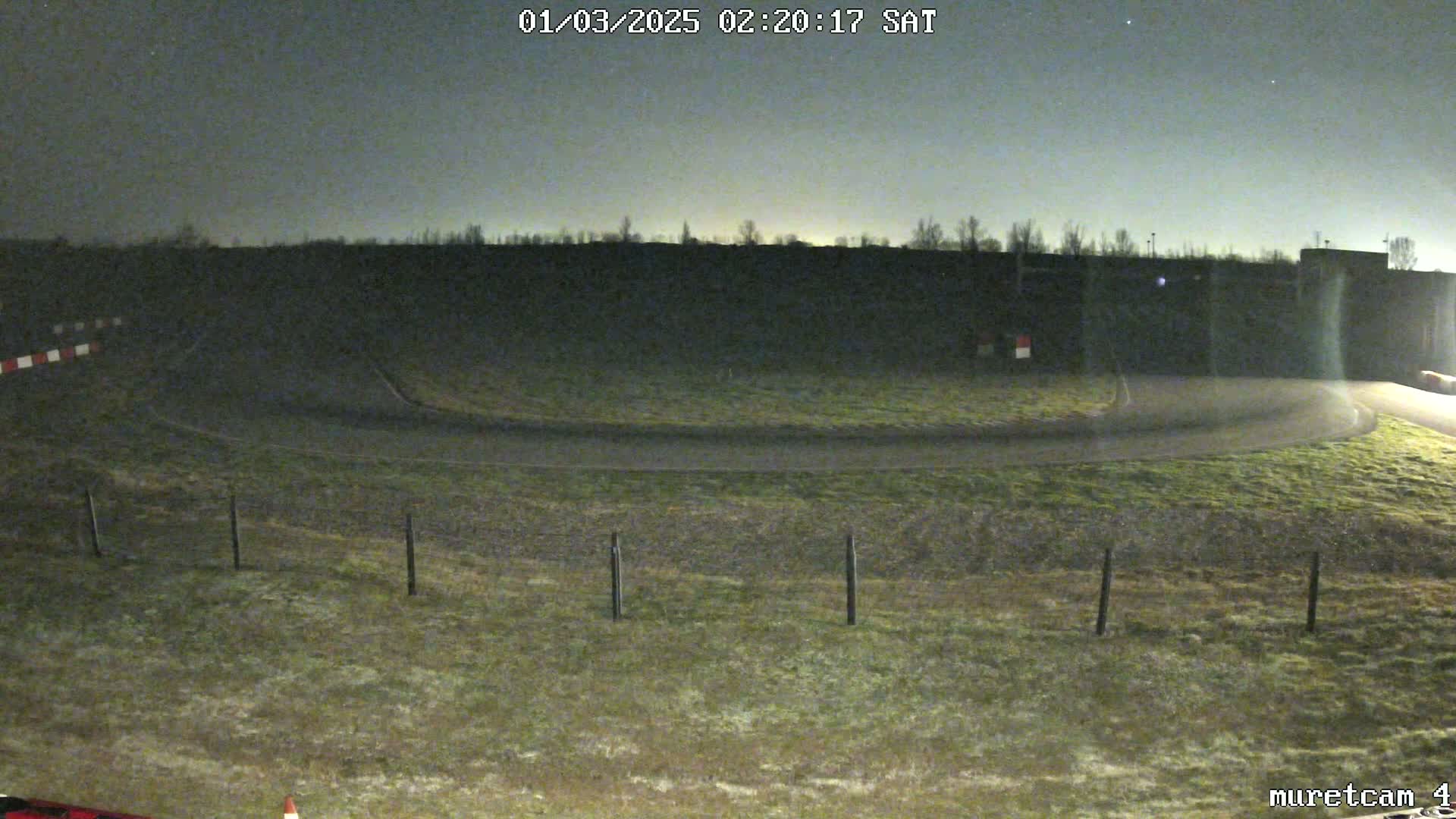 A nighttime view of a curved roadway with grassy areas and a fence, under a mostly clear sky.