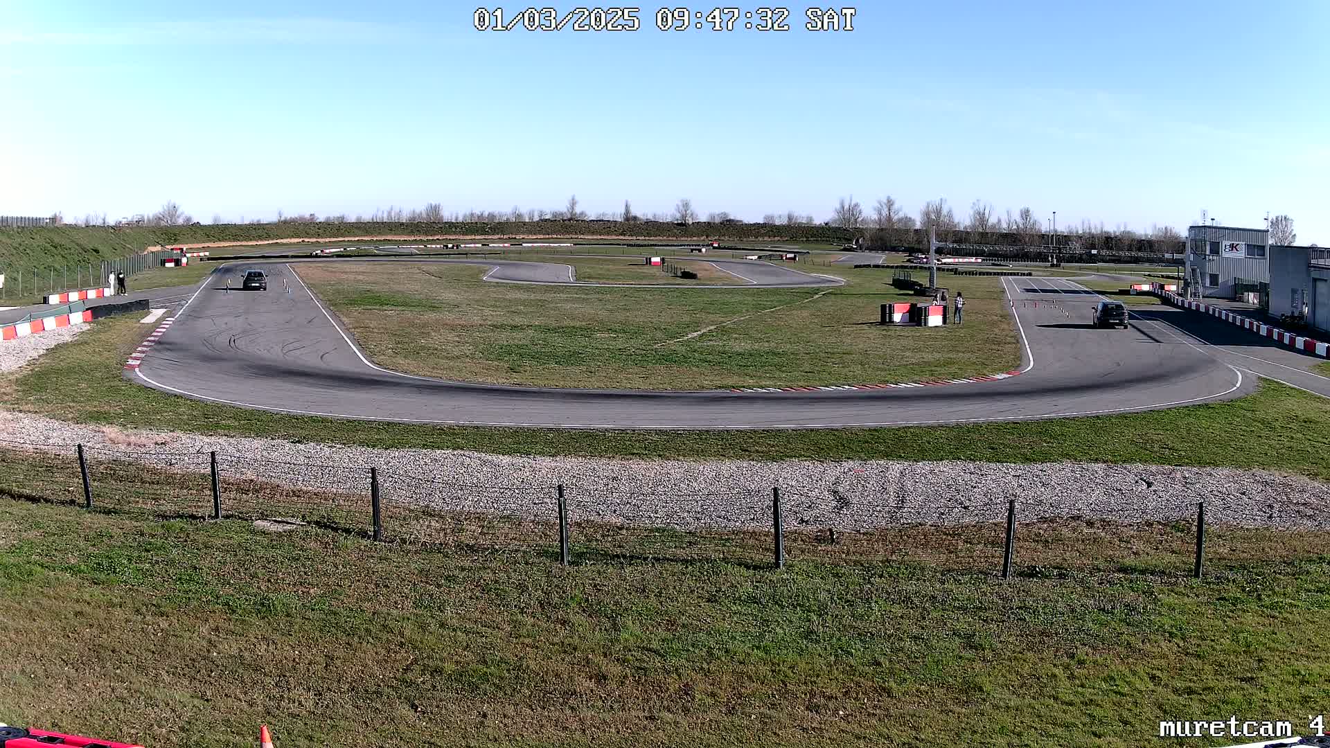 Under a clear blue sky, two cars are driving on a paved asphalt race track surrounded by grassy areas and a building in the background.