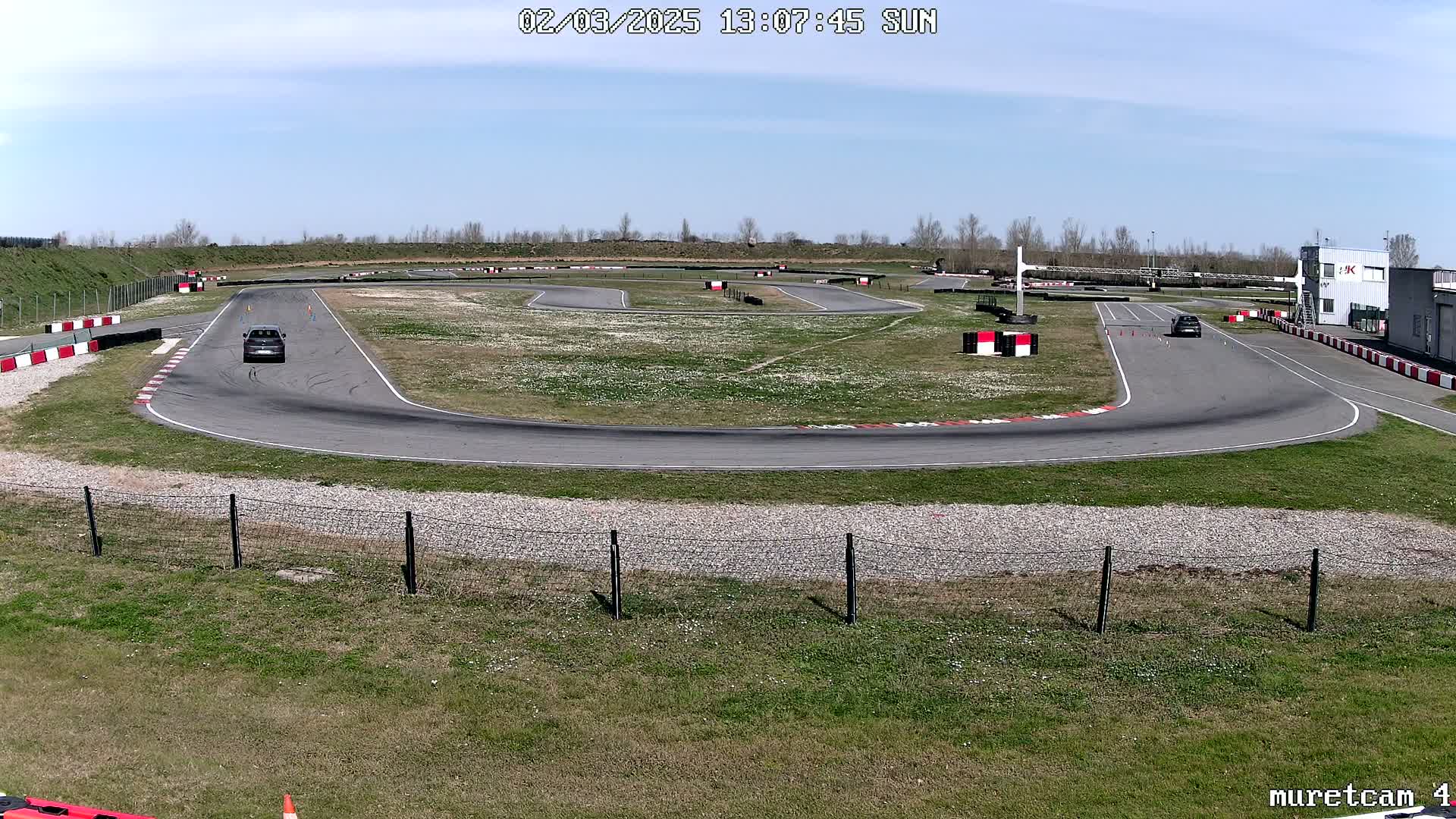 Two cars are driving on an asphalt race track surrounded by grassy areas under a sunny sky.