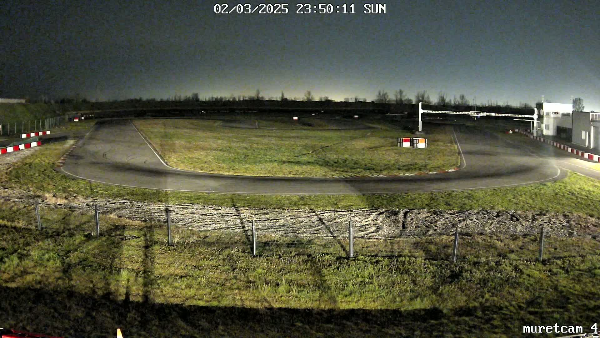 An empty asphalt go-kart track at night, surrounded by patchy grass and under a dark sky.