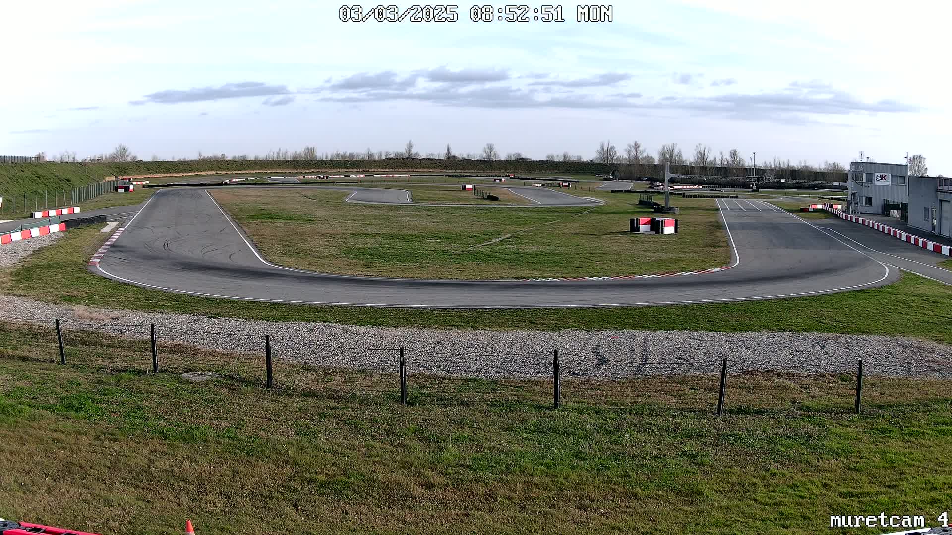 An outdoor go-kart track with a mostly sunny sky is visible, showing a winding asphalt course surrounded by grass and a building in the background.