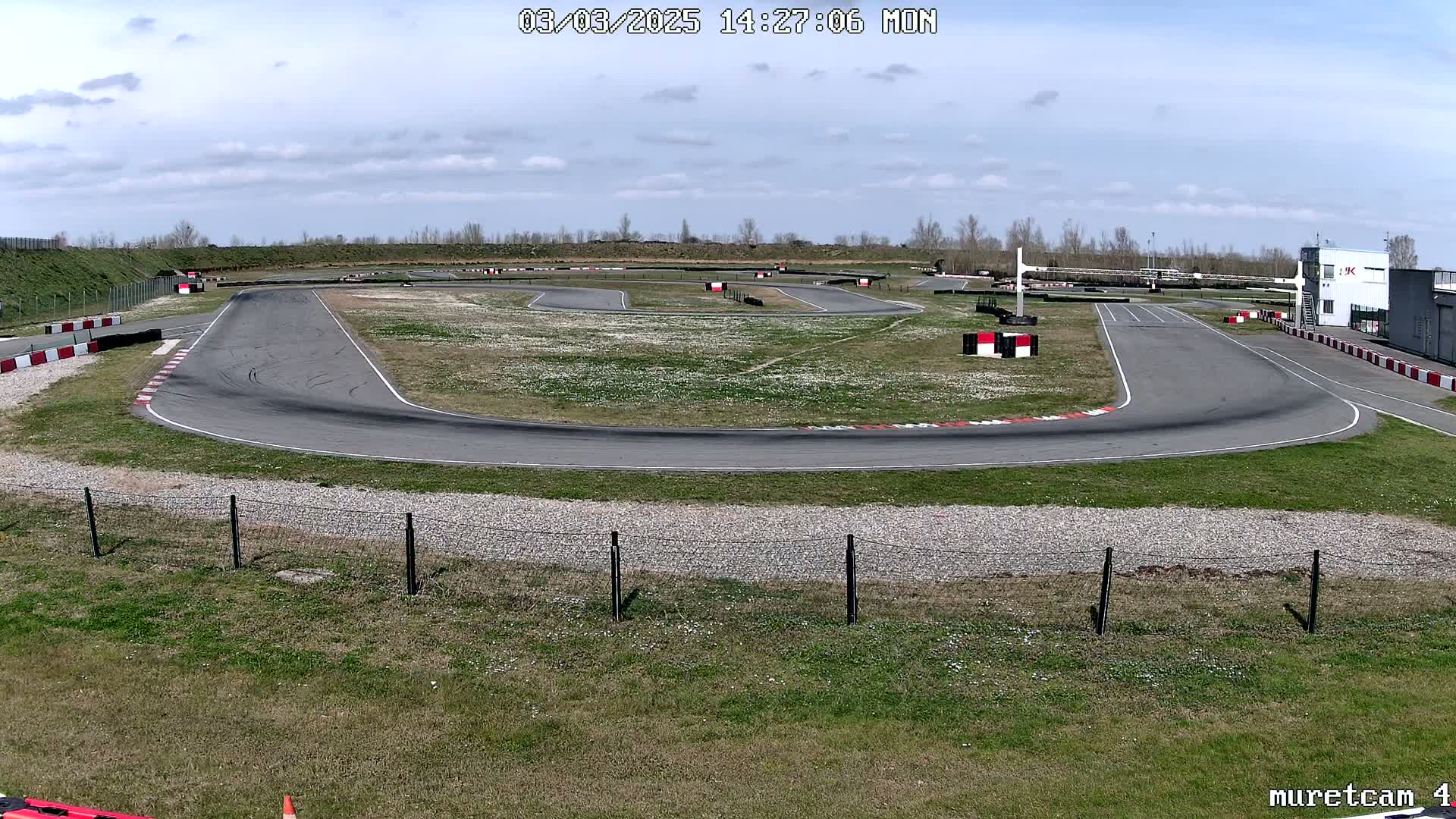 An empty asphalt go-kart track with patchy grass and wildflowers under a partly cloudy sky.