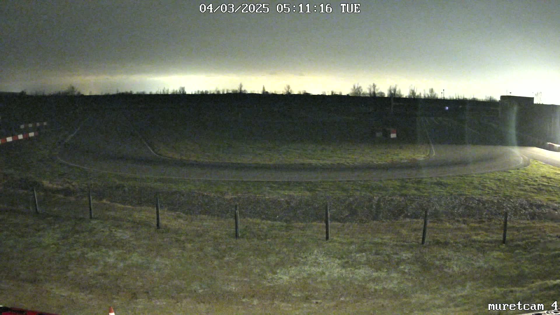 A dark, overcast night scene shows a paved, curved race track surrounded by short, grassy areas and a fence.