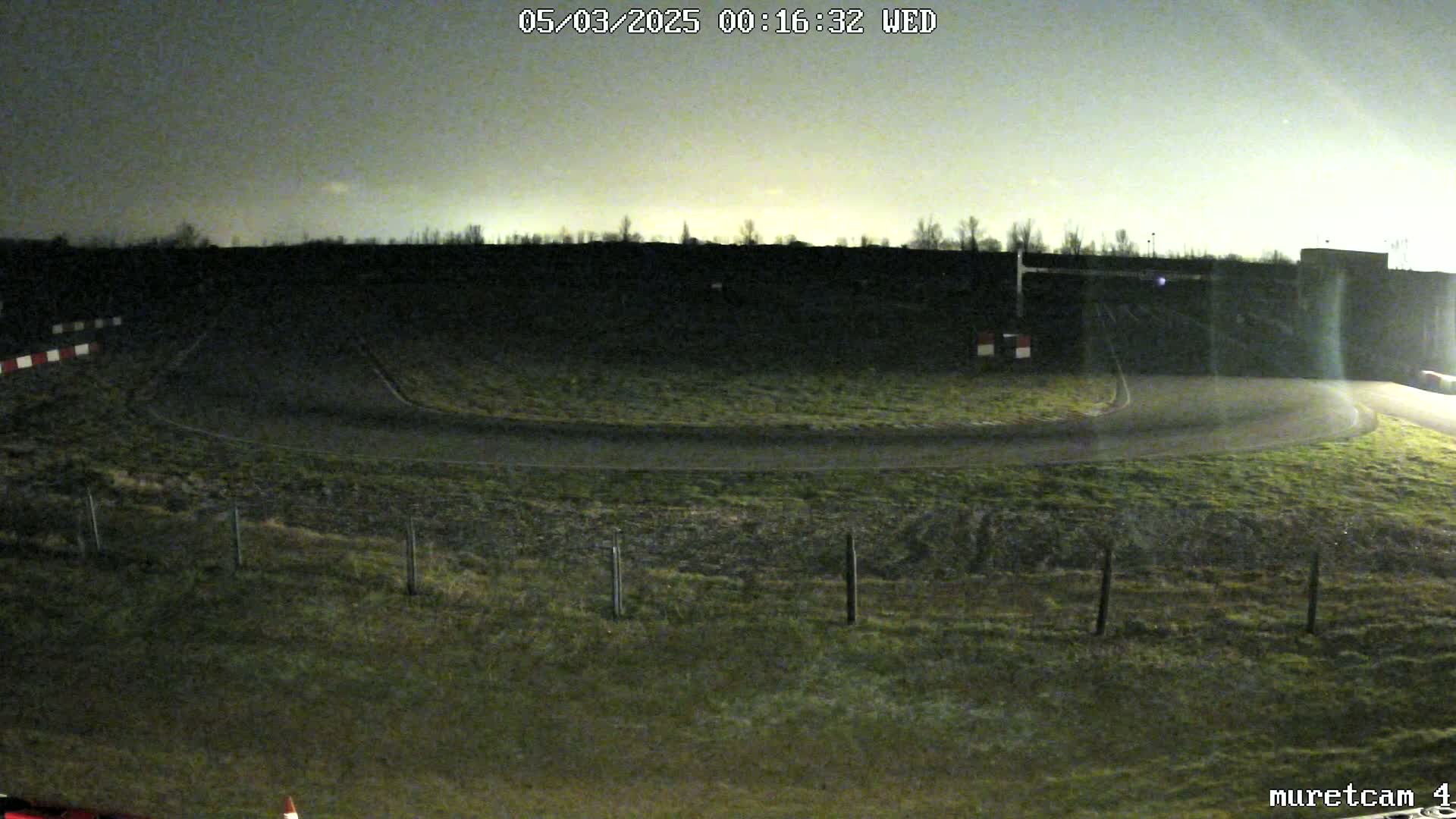 A nighttime view of a paved race track curving gently to the right, surrounded by short, dark-green grass under a mostly clear, dark sky.