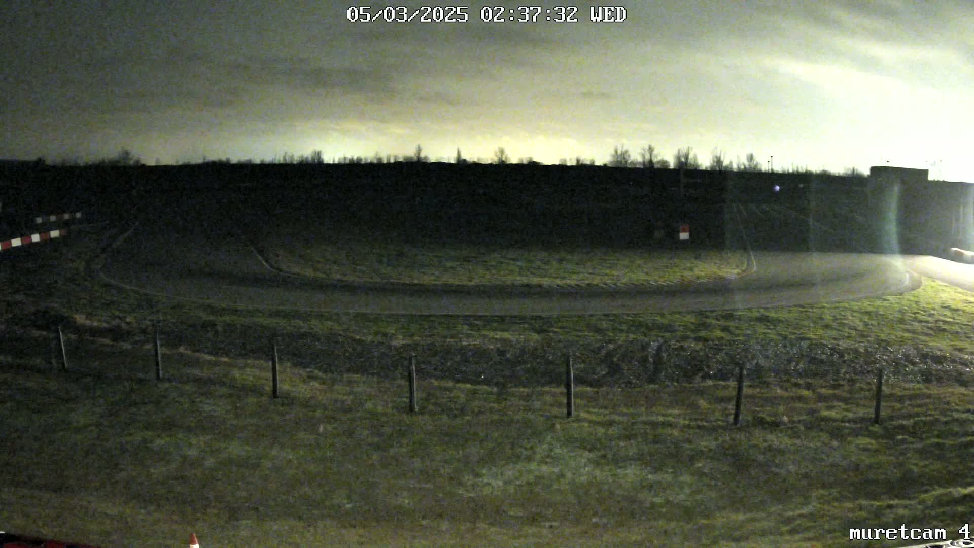 A nighttime, overcast view of a curved roadway bordered by grassy areas and a fence, with a dark wall in the background.