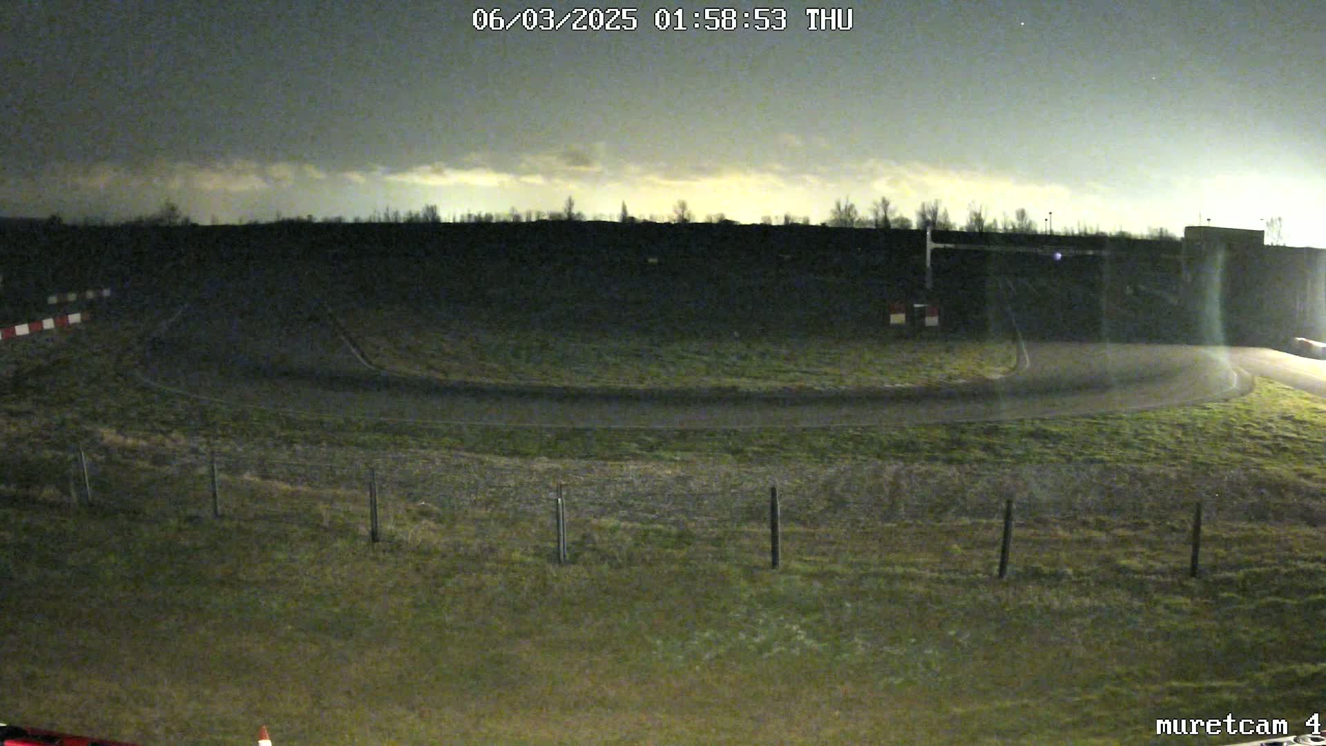 Under a partly cloudy night sky, a nighttime view shows a curved roadway with sparse vegetation and a fence bordering a grassy area.