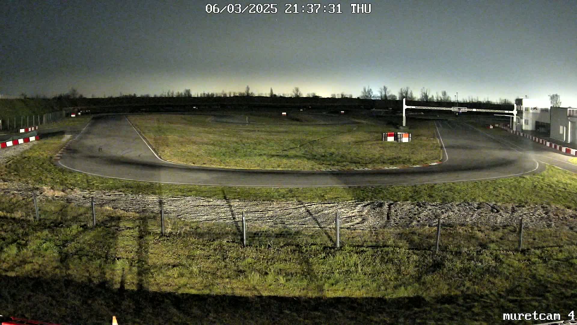 An empty asphalt go-kart track under a dark night sky is surrounded by short, grassy areas and fencing.