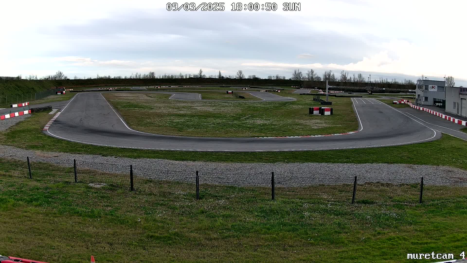 An empty asphalt go-kart track with red and white barriers curves through a grassy field under a cloudy sky.