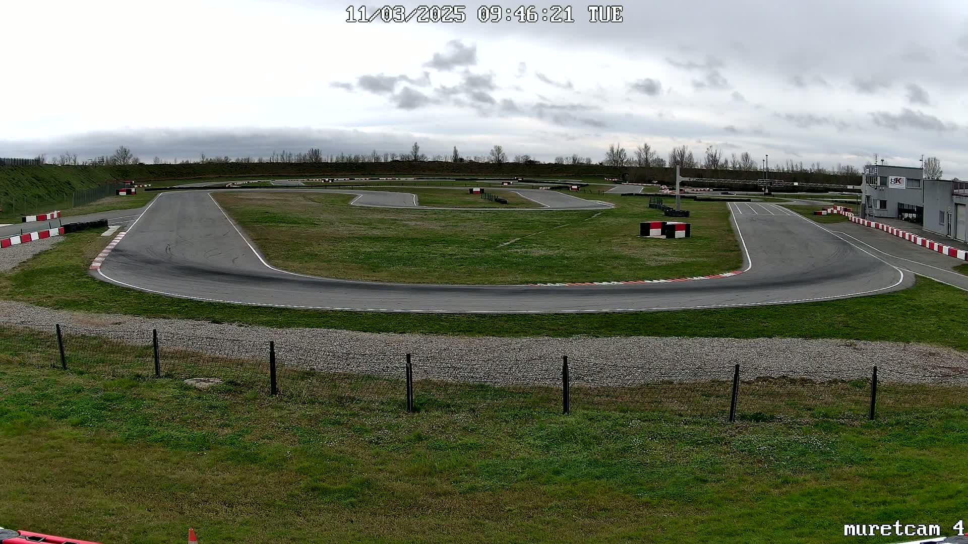 An outdoor go-kart track with overcast skies curves around a grassy area, bordered by a fence.