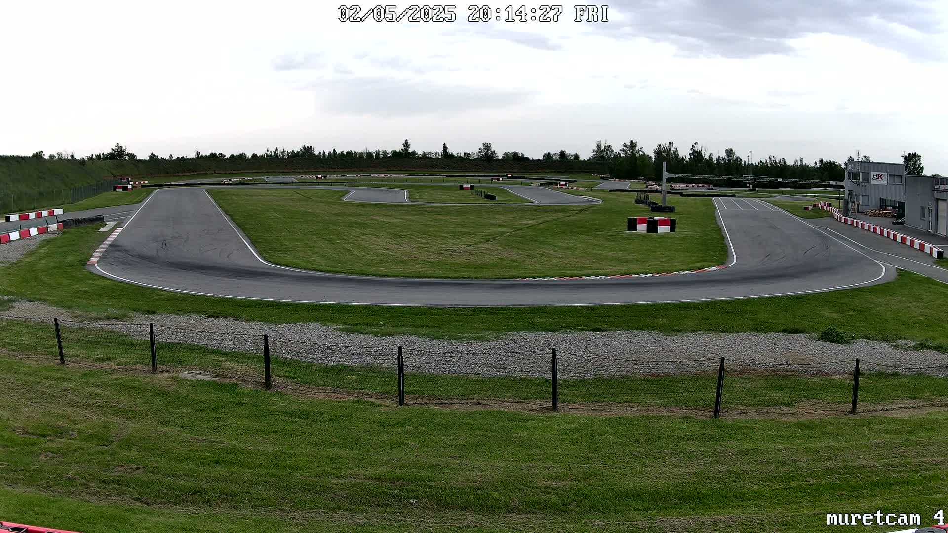 An outdoor go-kart track with overcast skies is surrounded by grassy areas and a chain-link fence.