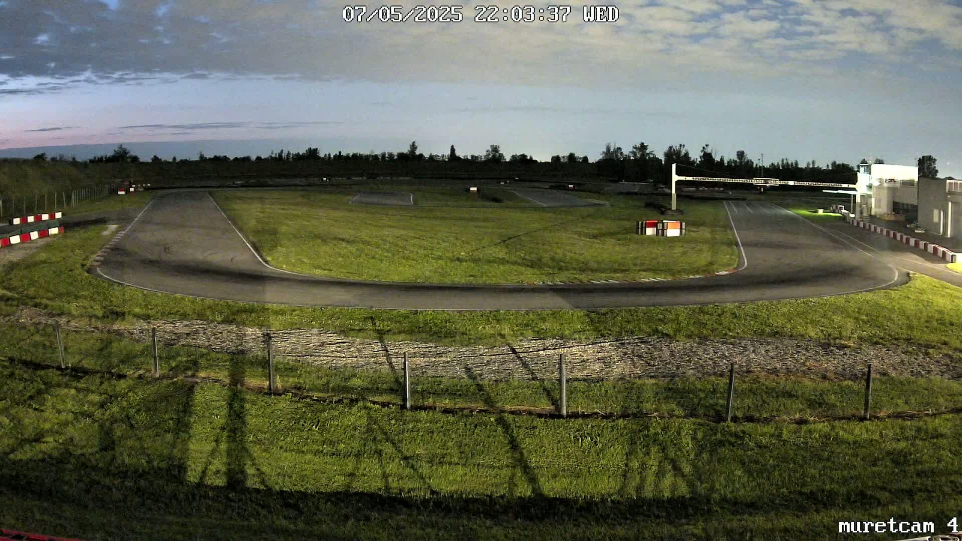 An empty go-kart track under a twilight sky is surrounded by grassy areas and a low fence.