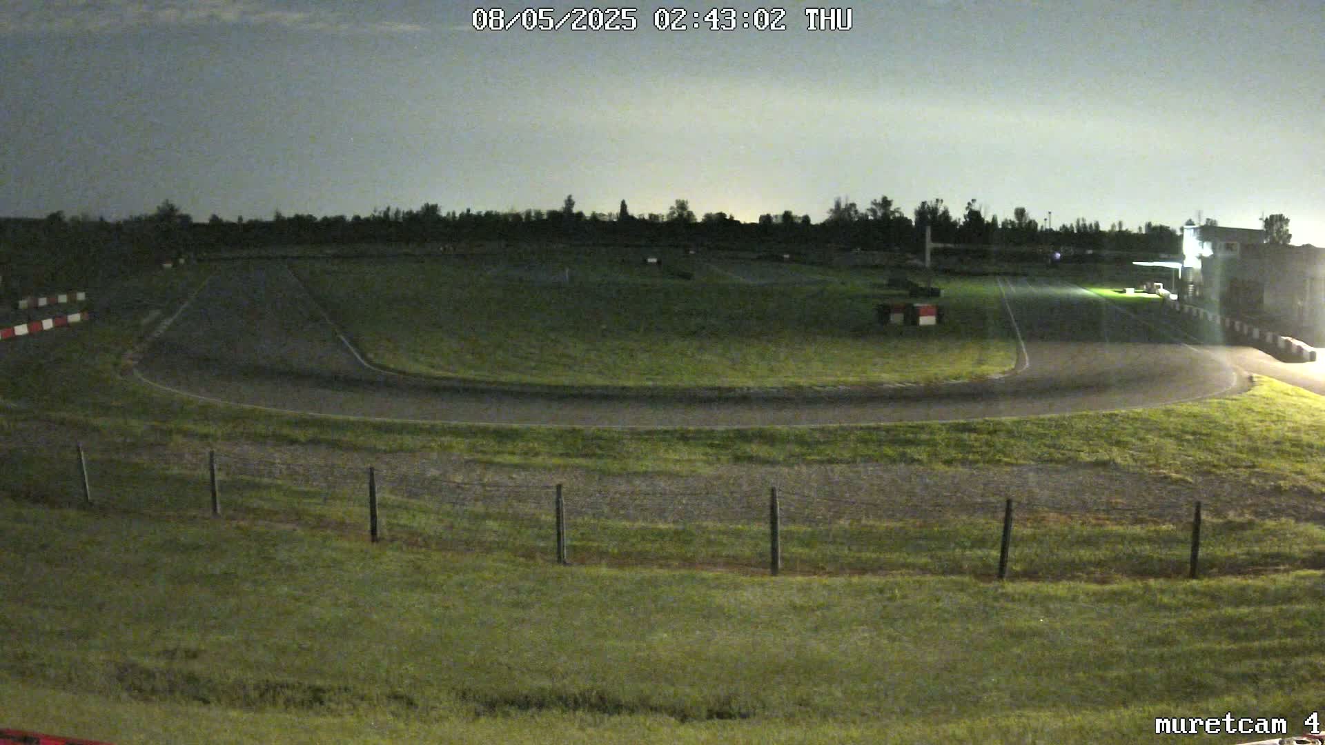 A nighttime view of a go-kart track, mostly empty and surrounded by grass under a cloudy sky.