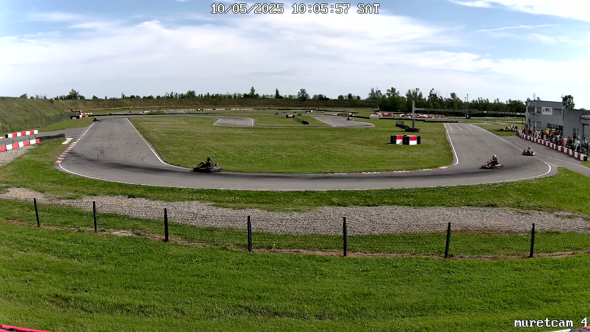 Two go-karts race around a paved outdoor track on a partly cloudy day.