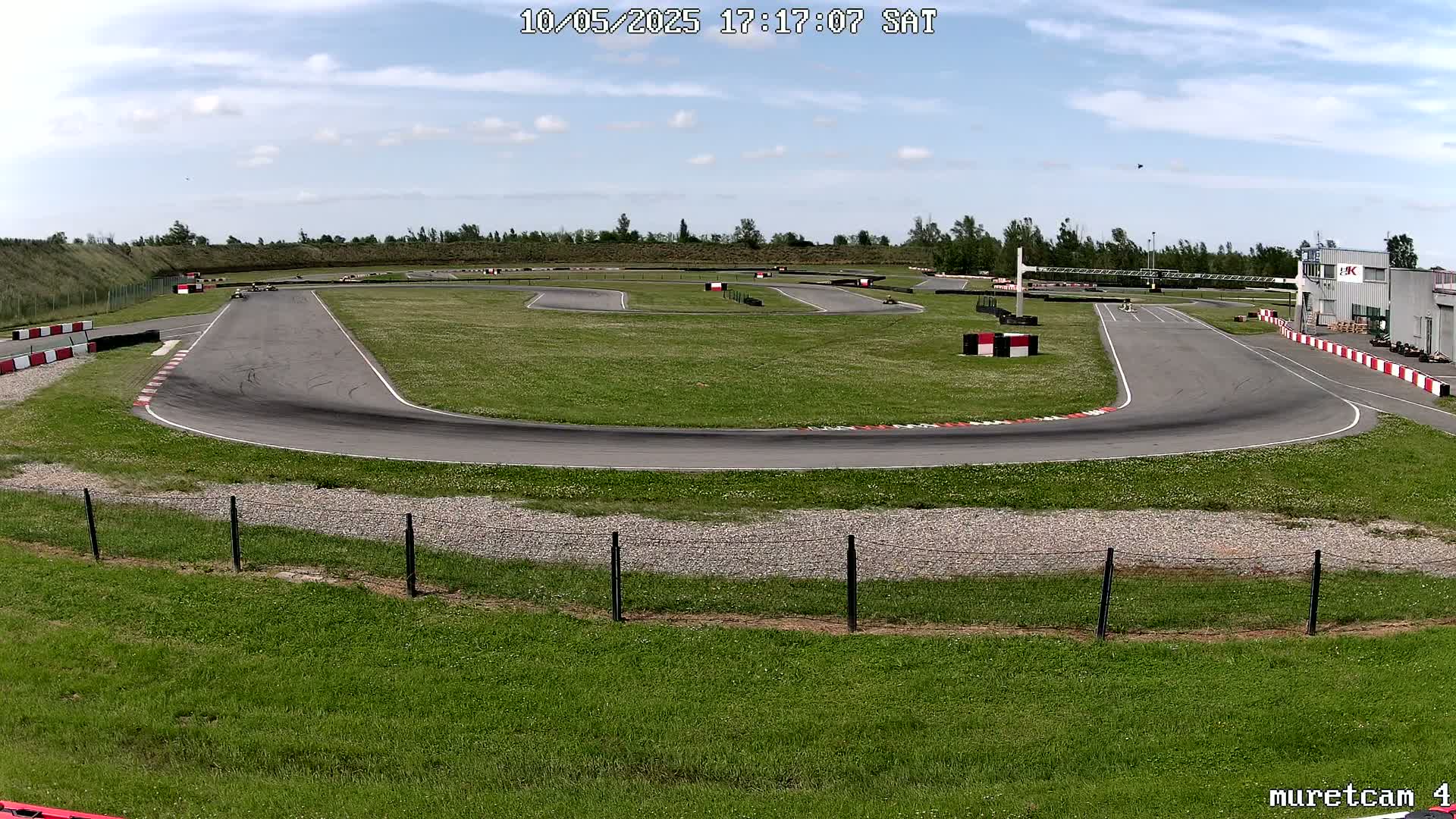 An outdoor go-kart track on a partly sunny day curves through a grassy field, with a few go-karts visible on the track.