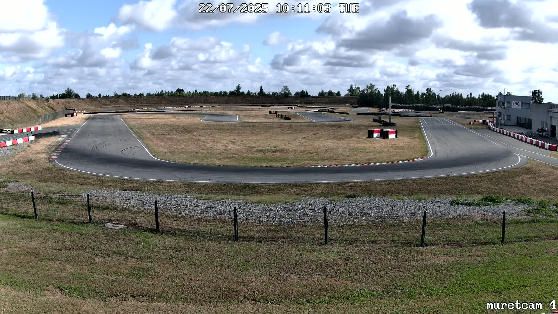 An outdoor go-kart track with a partly cloudy sky, under sunny conditions, is surrounded by dry grass and a chain-link fence.