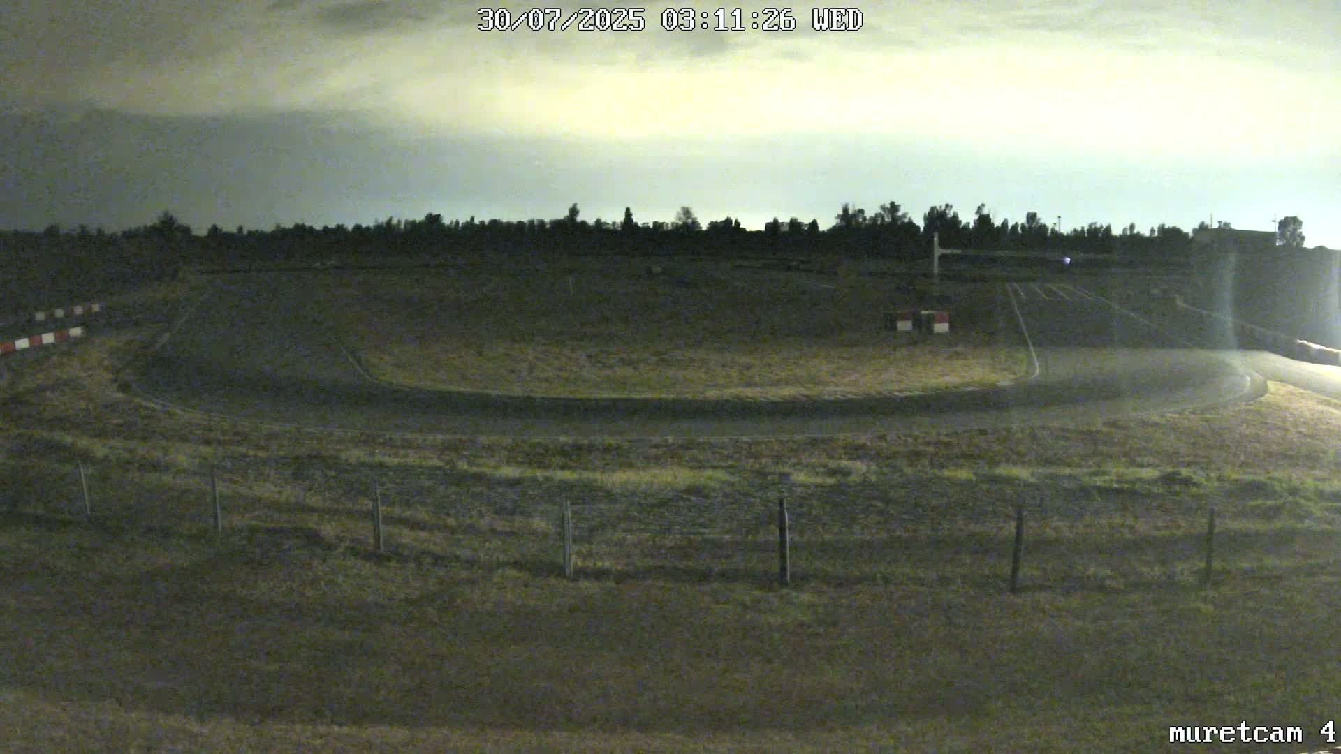 An outdoor go-kart track with a partly cloudy sky, under sunny conditions, is surrounded by dry grass and a chain-link fence.