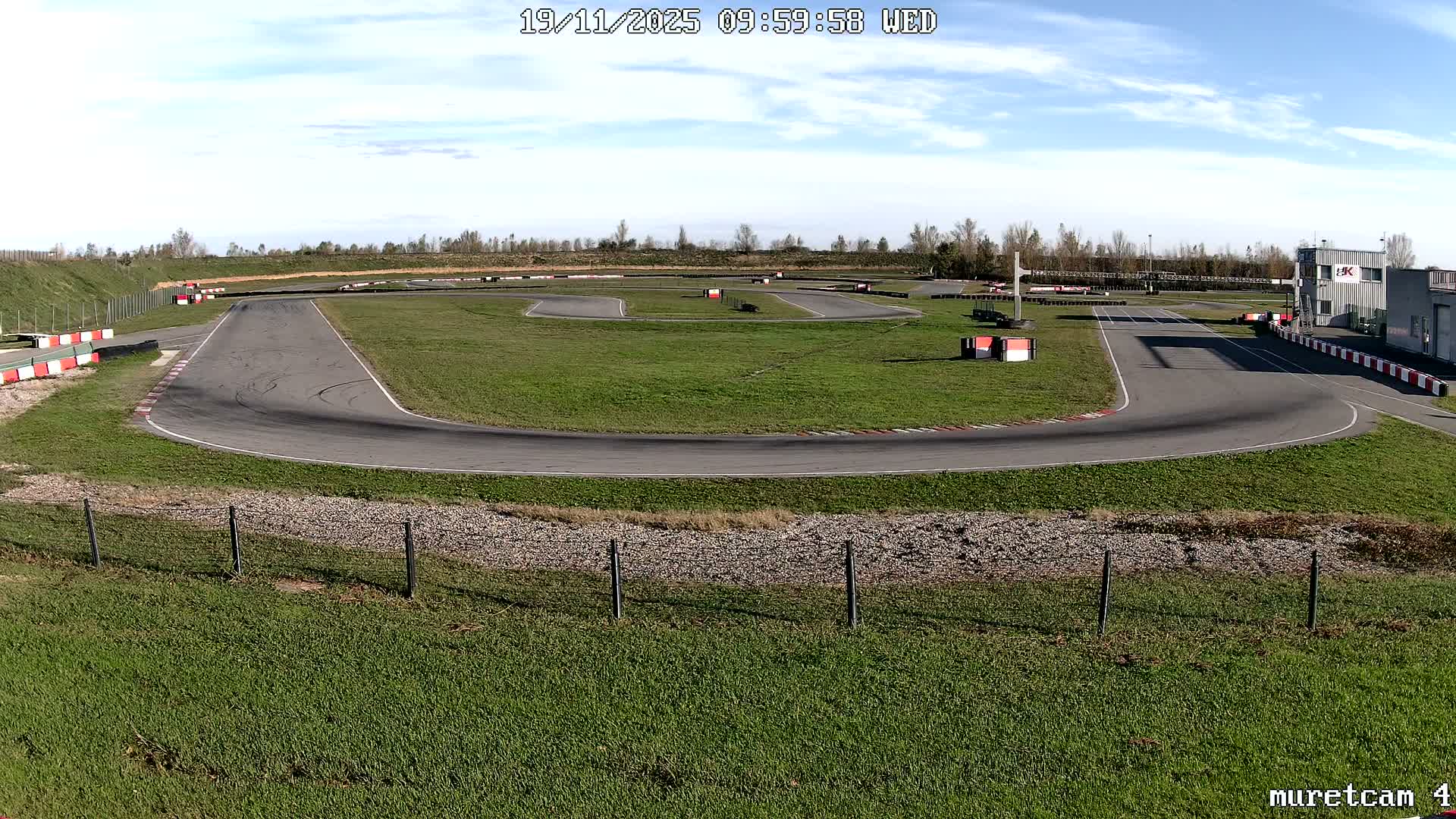 An empty outdoor karting track with green grassy verges, several turns, barriers, and a small building in the background is seen under a partly cloudy sky.