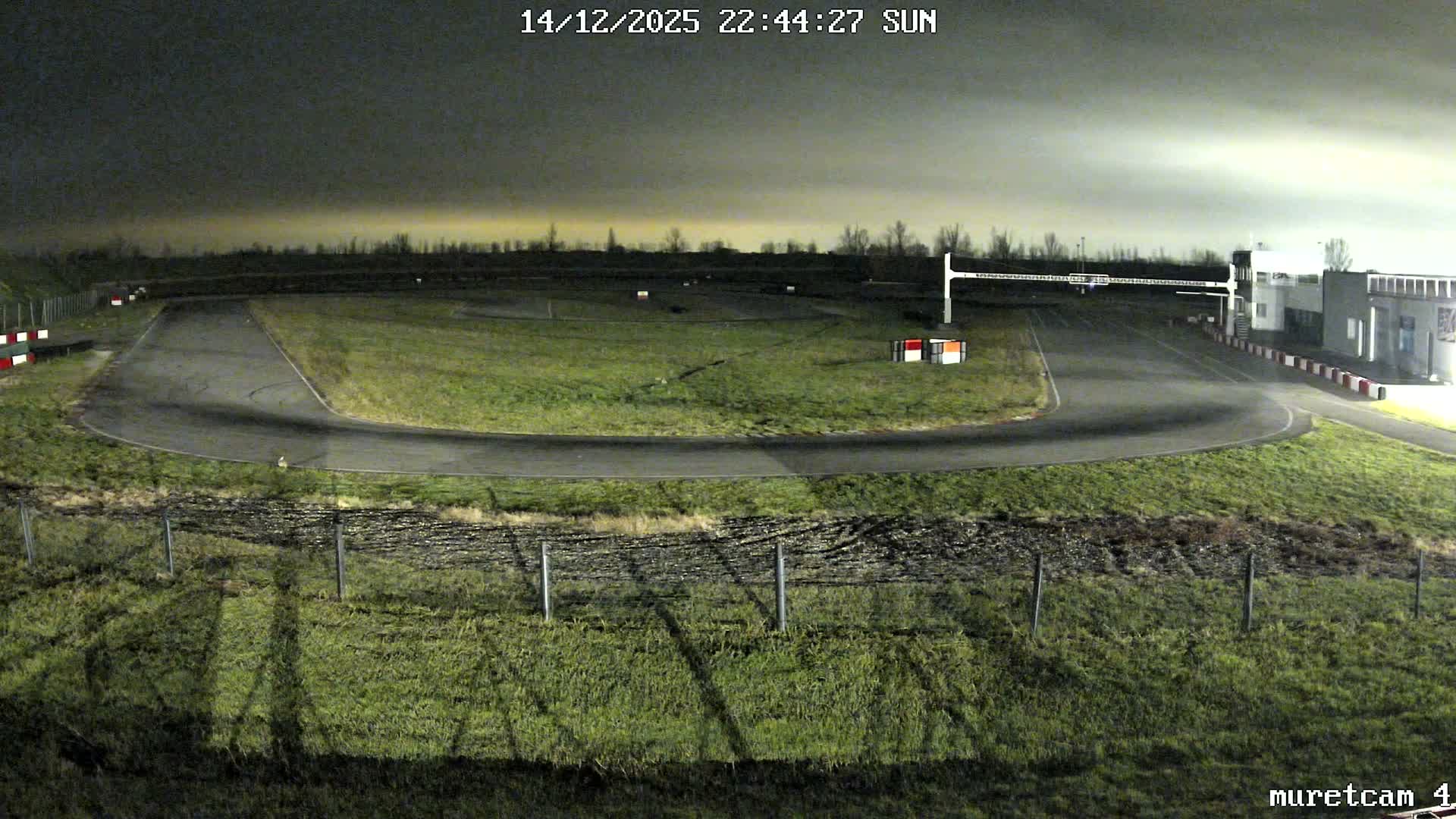 A go-kart track with several cars and a building is visible under a grey, overcast sky, showing damp ground and some puddles.
