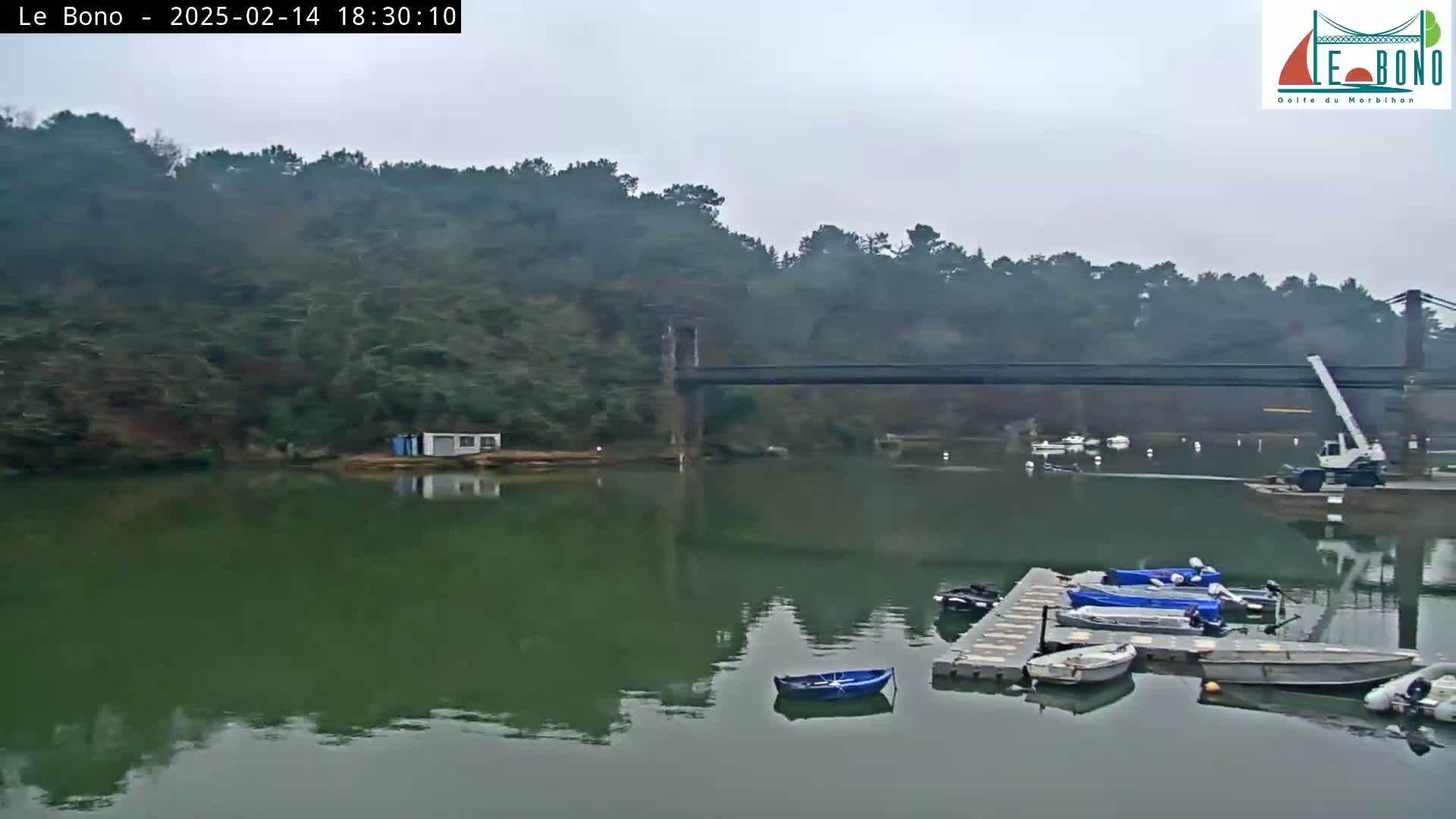 A calm, overcast day shows a suspension bridge spanning a river with several small boats moored at a dock near a wooded hillside.