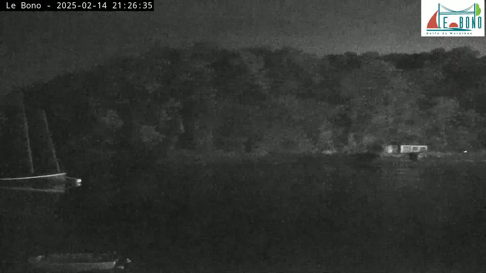 A nighttime, low-light image shows a dark body of water with a boat on the left and a dark shoreline with trees and a small structure on the right.