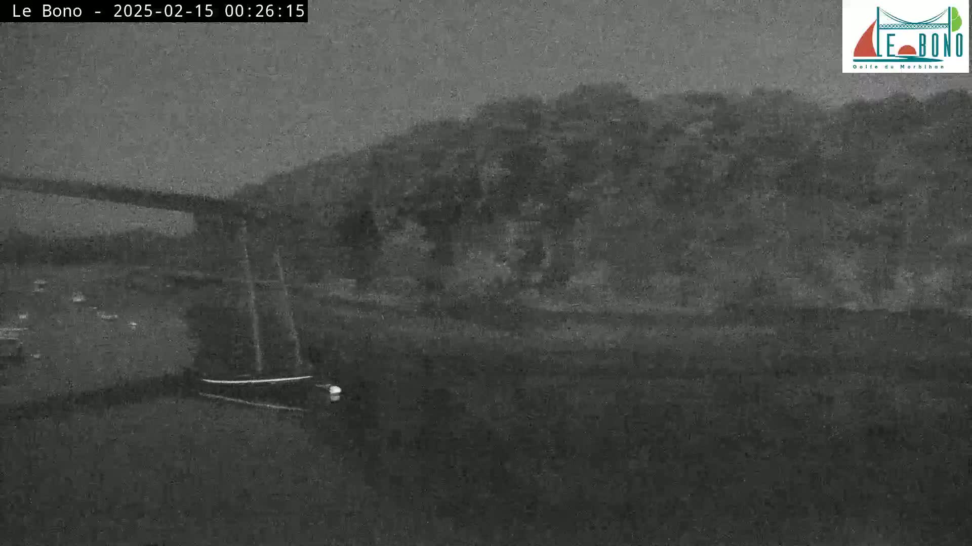 A nighttime, low-light image shows a dark body of water with a small boat and a dark shoreline with trees under a cloudy sky.