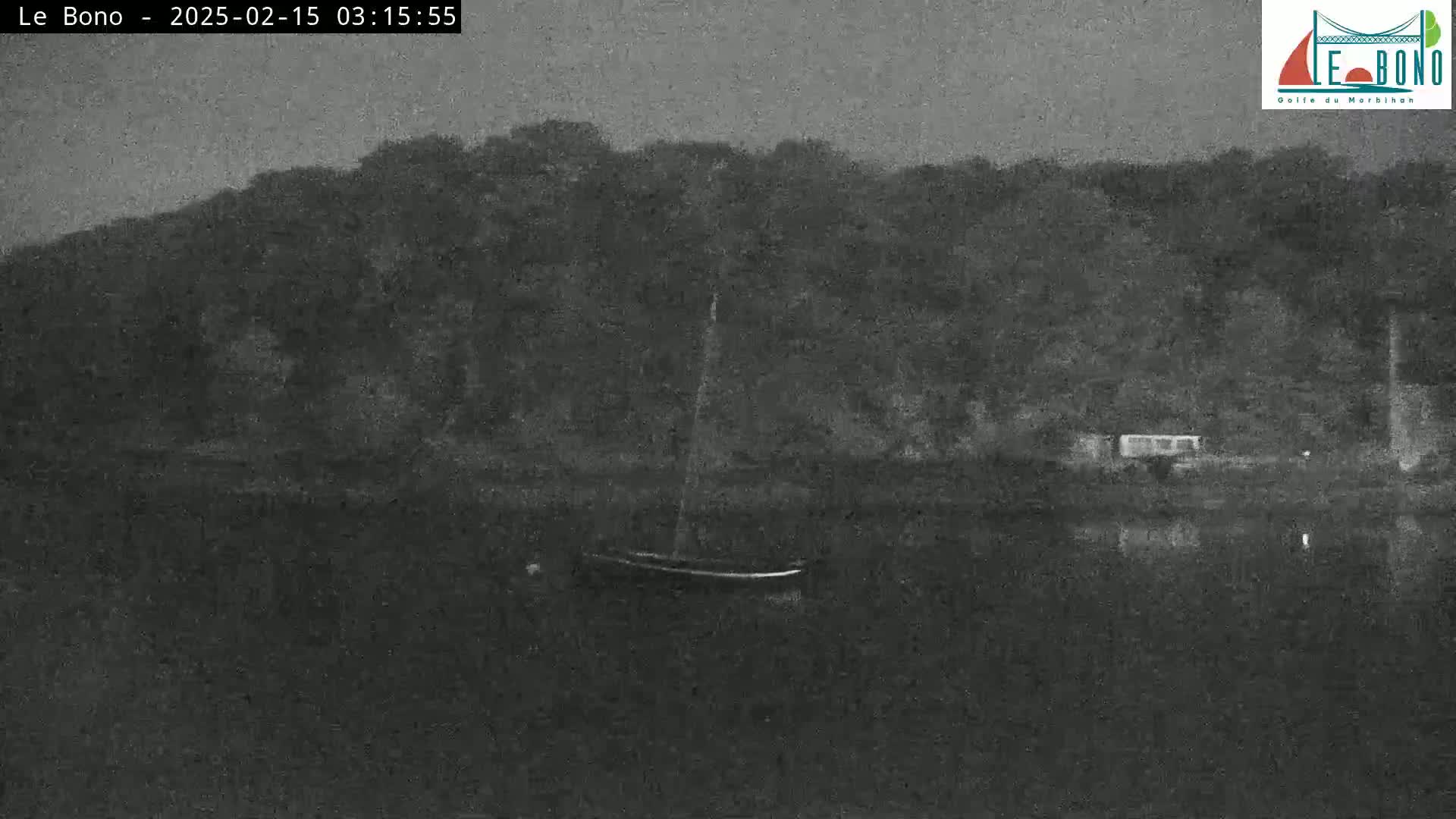 A small sailboat is moored in dark, still water in front of a dark, wooded shoreline under a dim, nighttime sky.