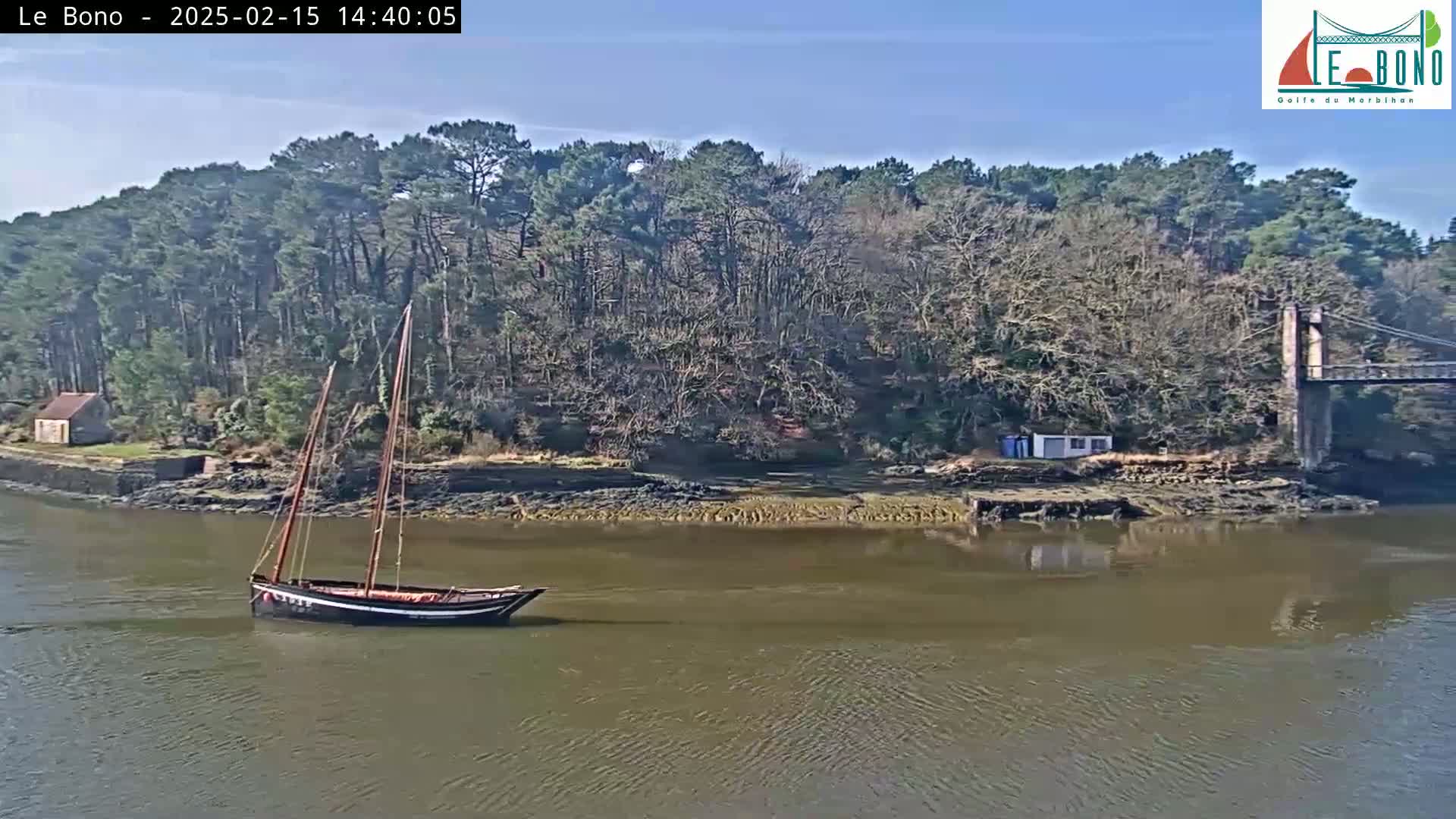 A dark-colored sailboat is in a calm body of water near a wooded shoreline and a suspension bridge under a sunny sky.