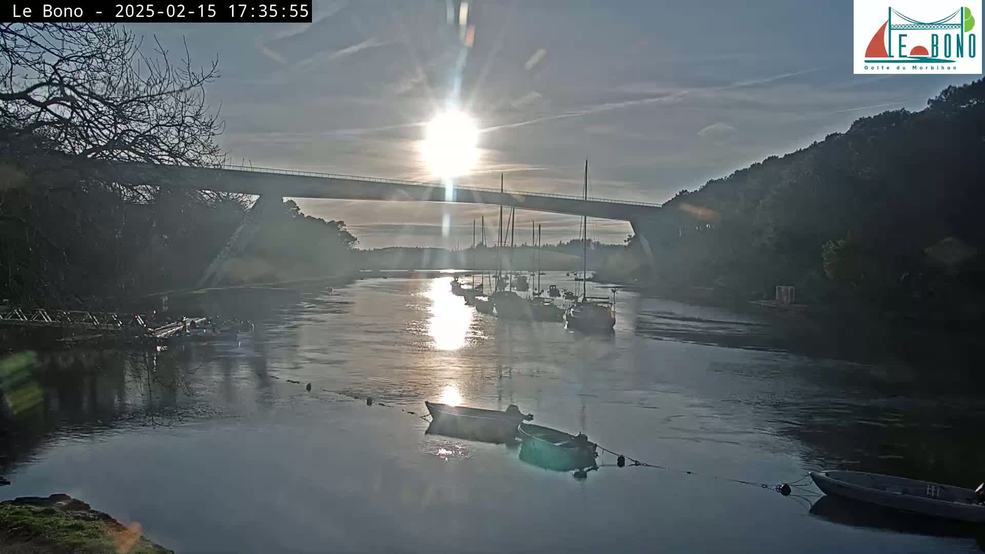 A calm river at sunset, with several boats moored beneath a long bridge, and trees lining the banks.