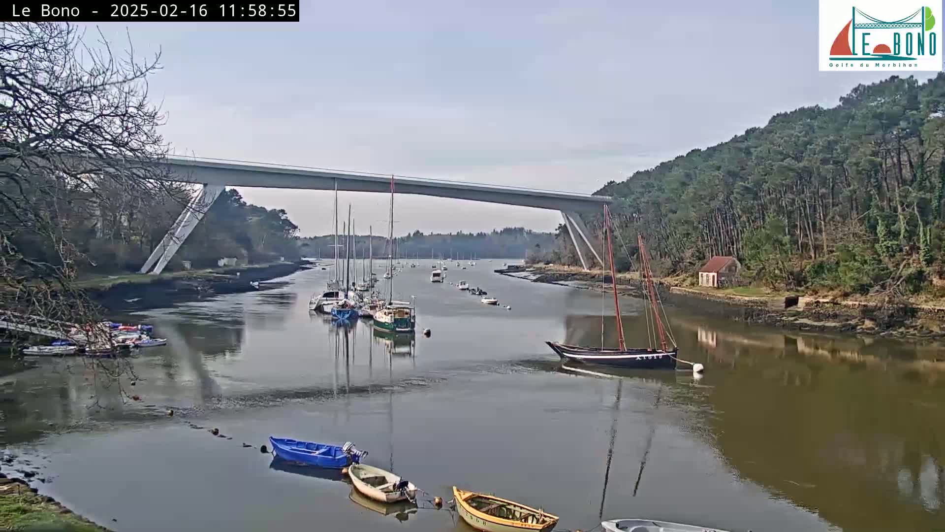 Several sailboats are moored in a calm river under a long bridge on a partly sunny day.