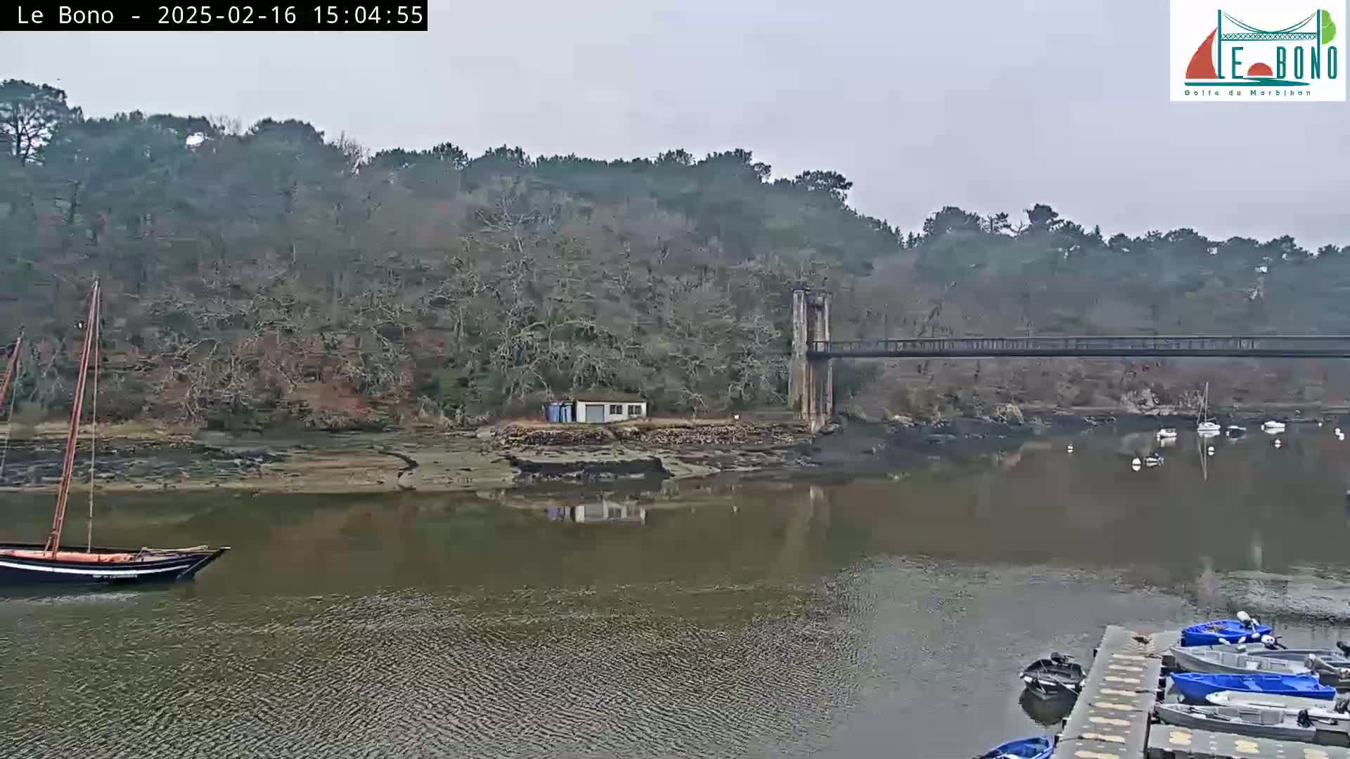 A river scene on an overcast day shows a small sailboat, a footbridge spanning the river, several small motorboats docked, and a wooded hillside in the background.
