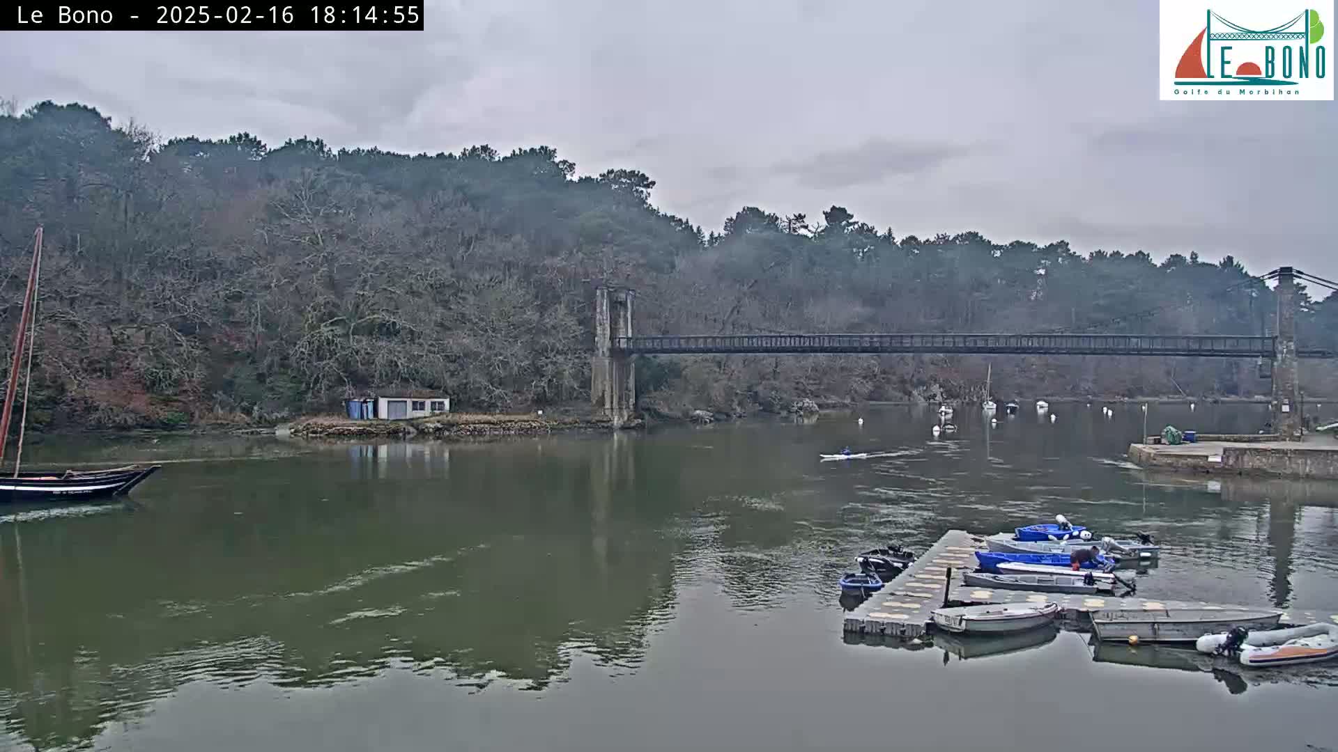 A suspension bridge spans a calm river under an overcast sky, with several small boats moored at a dock and a sailboat in the distance.