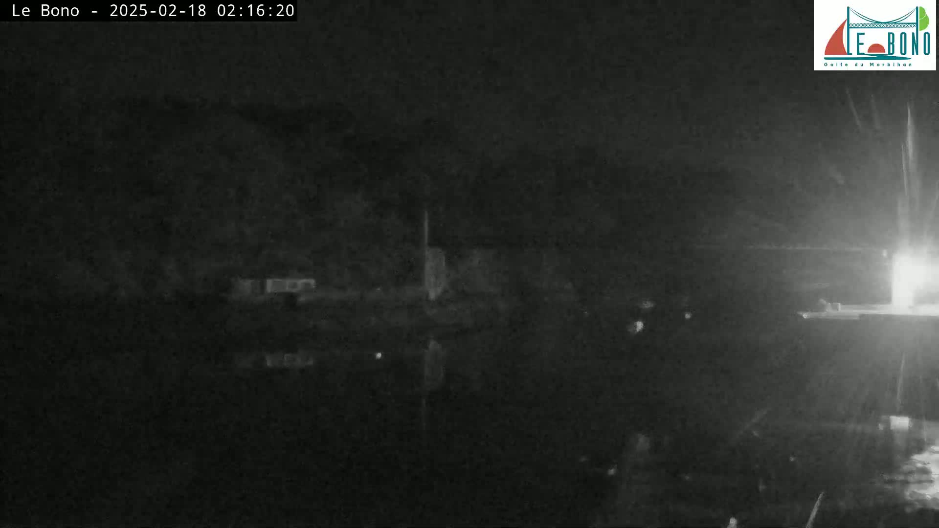 A nighttime, low-light image shows a dark body of water reflecting a dark shoreline under a seemingly rainy sky.