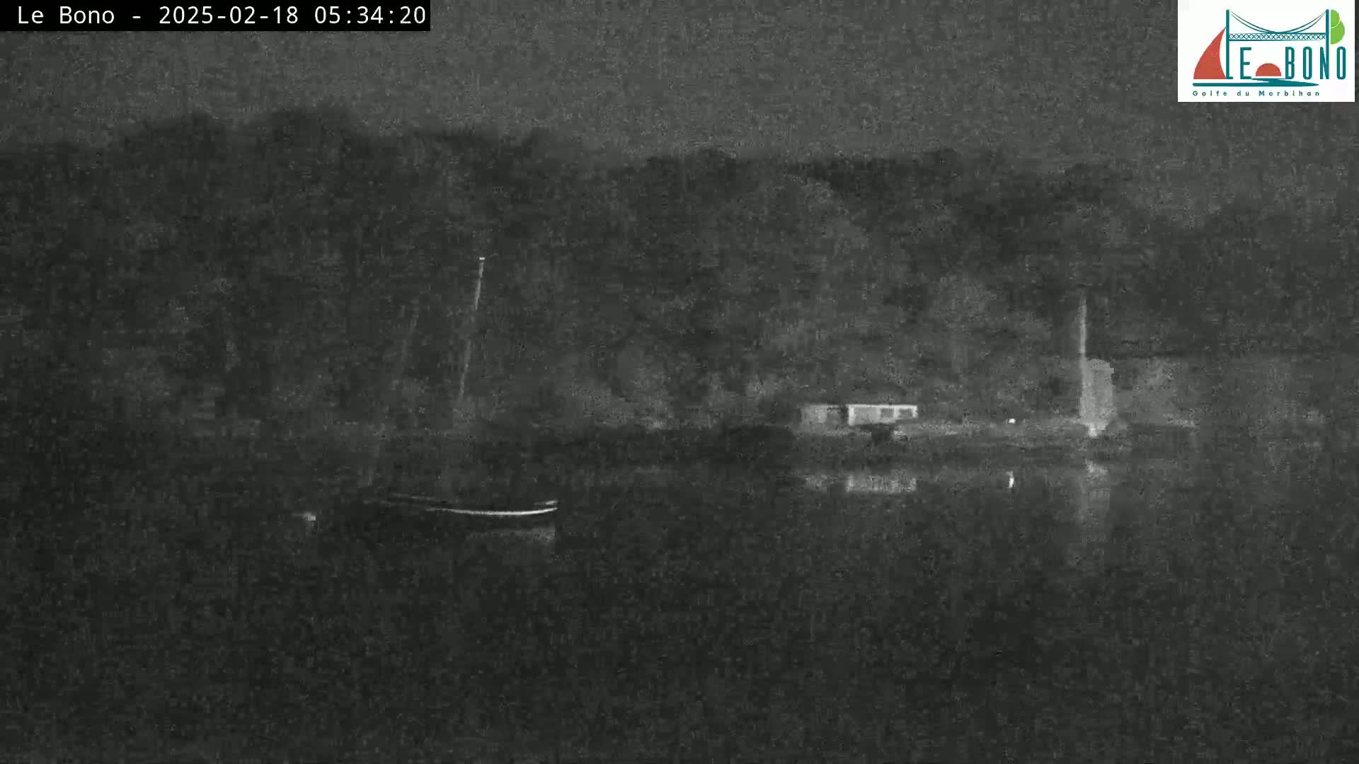 A nighttime, low-light image shows a dark body of water reflecting the shadowy outline of trees and buildings on the far shore.