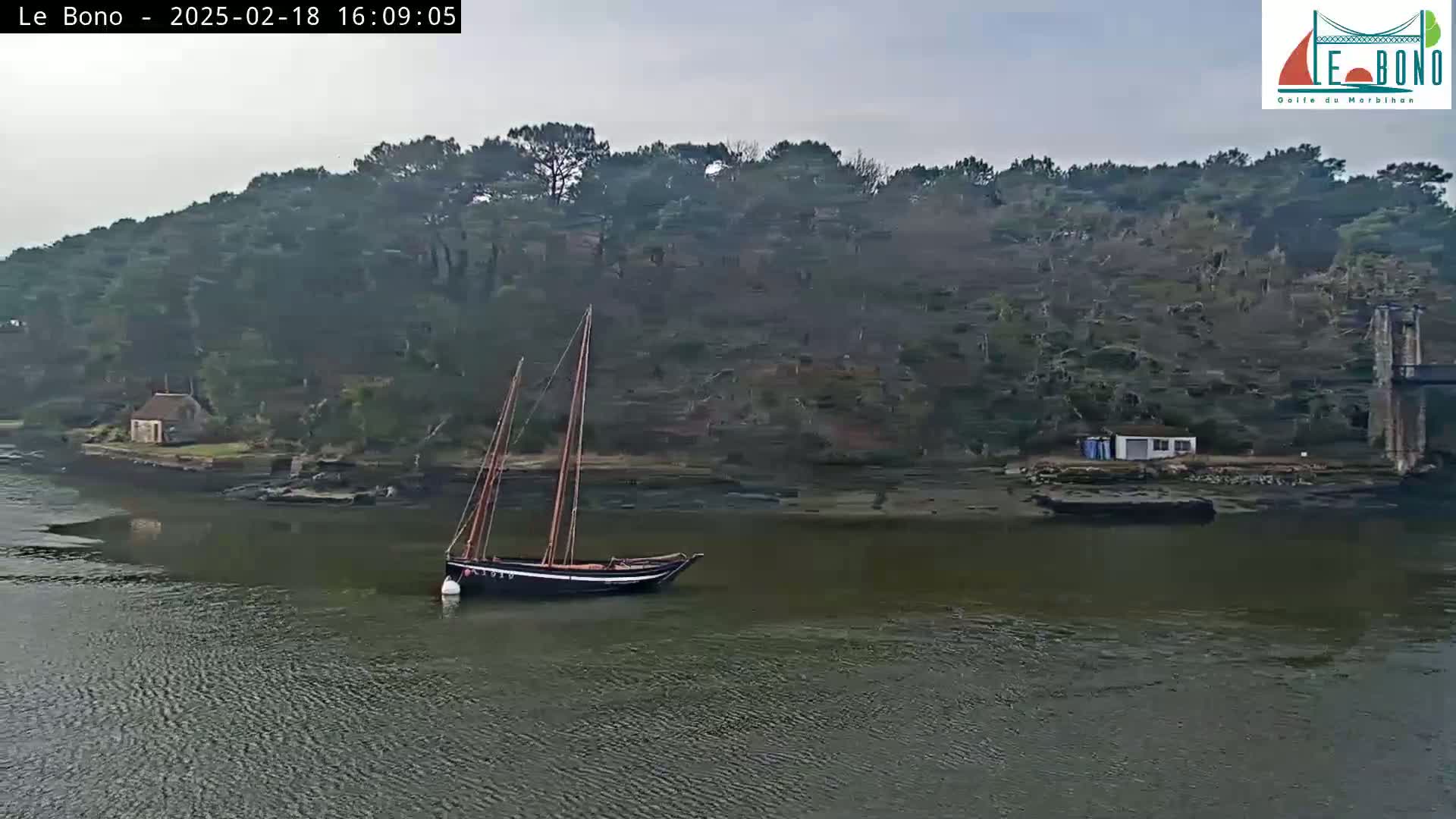 A dark-hulled sailboat is moored in calm, gray water near a shoreline with dark green trees and a small white building under an overcast sky.
