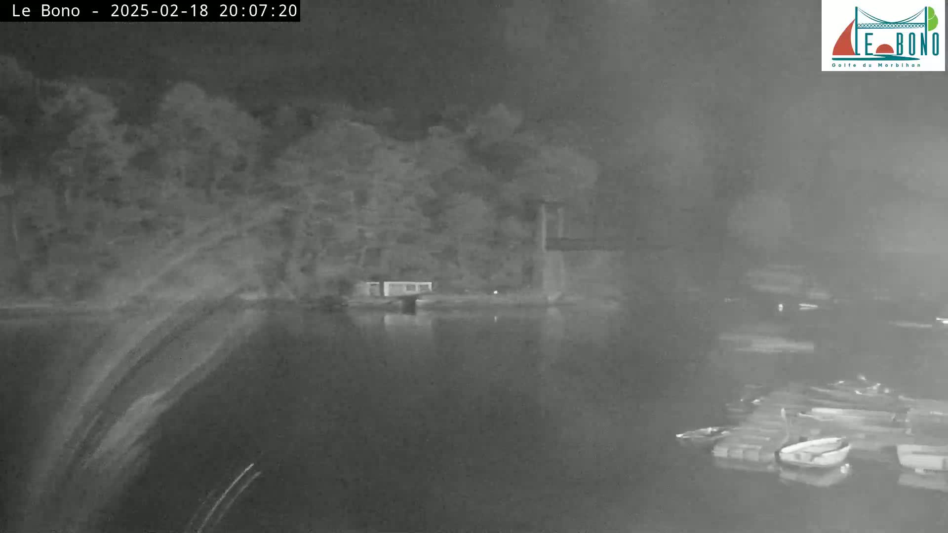A nighttime, low-light image shows a calm body of water with several small boats moored near a shoreline with trees and a small building under a dark, possibly cloudy sky.