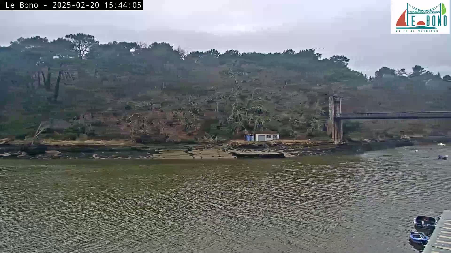 A calm body of water with a few small boats near a shoreline featuring a small building and a suspension bridge behind a tree-covered hill under an overcast sky.