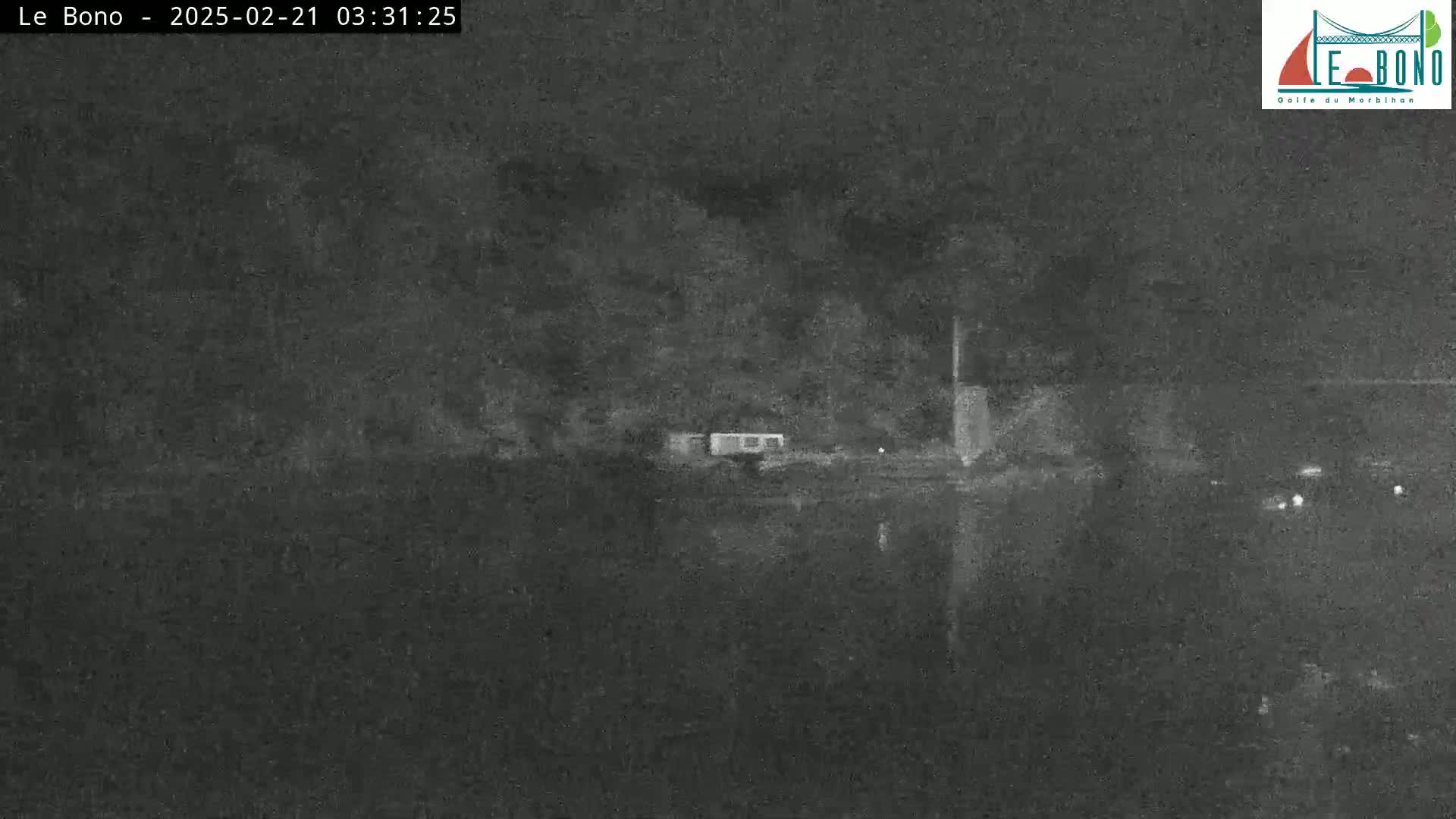 A nighttime, low-light image shows a dark body of water reflecting a dark shoreline with some structures and vegetation.