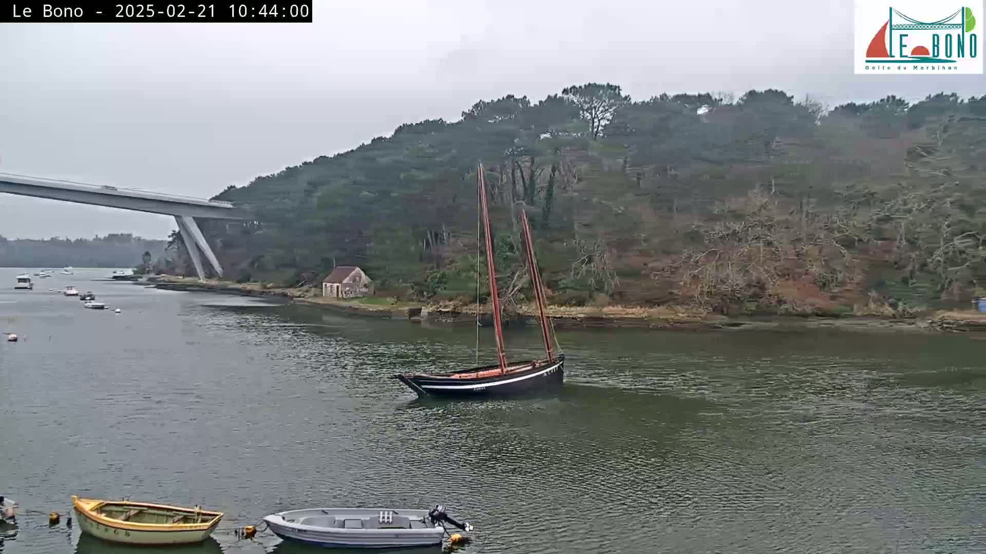 A sailboat is moored in a calm waterway under an overcast sky, near a bridge and a wooded shoreline with several small boats nearby.