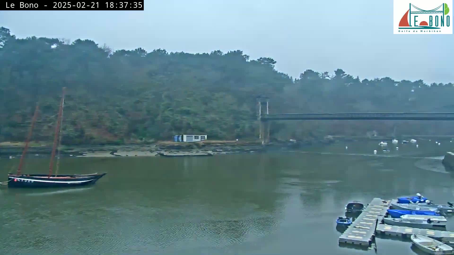 A sailboat is moored in a calm river under an overcast sky, with a bridge in the background and several small boats docked at a pier.