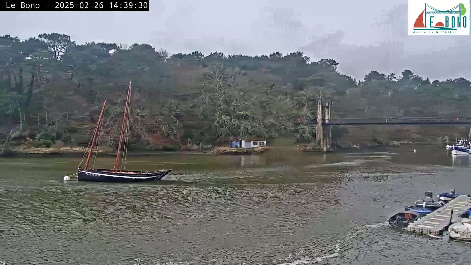 A dark-hulled sailboat is anchored in a calm, overcast body of water near a suspension bridge and several smaller boats moored at a dock, with a tree-covered hillside in the background.