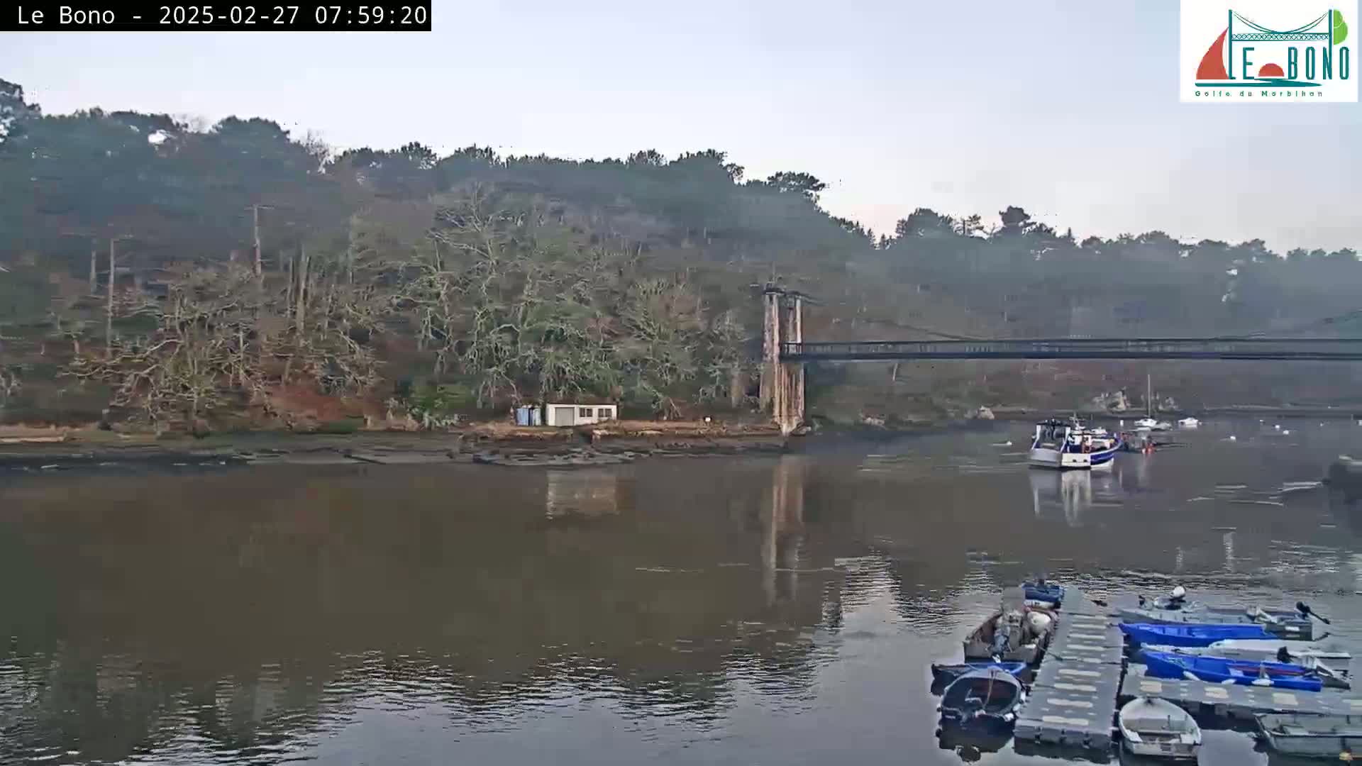 A calm river flows under a suspension bridge, past a small building on the bank, with several boats moored on a floating dock and others sailing on the river under an overcast sky.