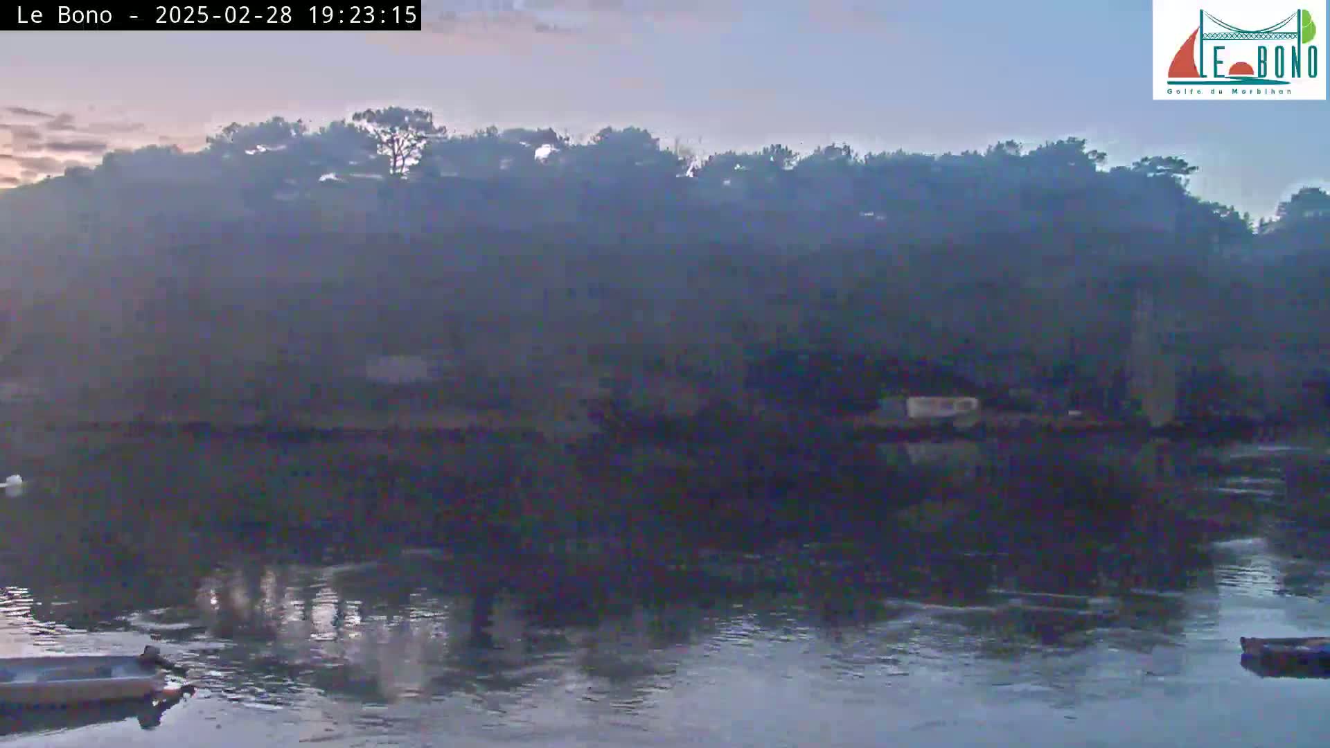 A calm body of water reflects a dusky sky at twilight, with a dark line of trees and low-lying buildings visible on the far shore.
