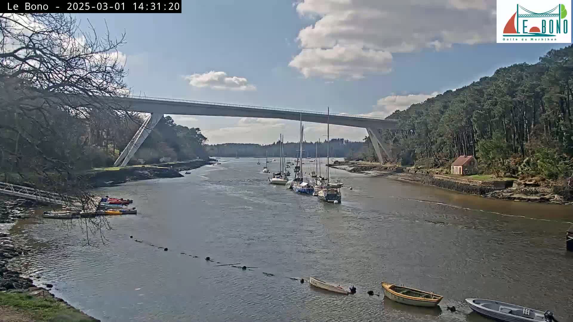 A long bridge spans a calm river where several sailboats and small motorboats are moored, under a partly cloudy sky.